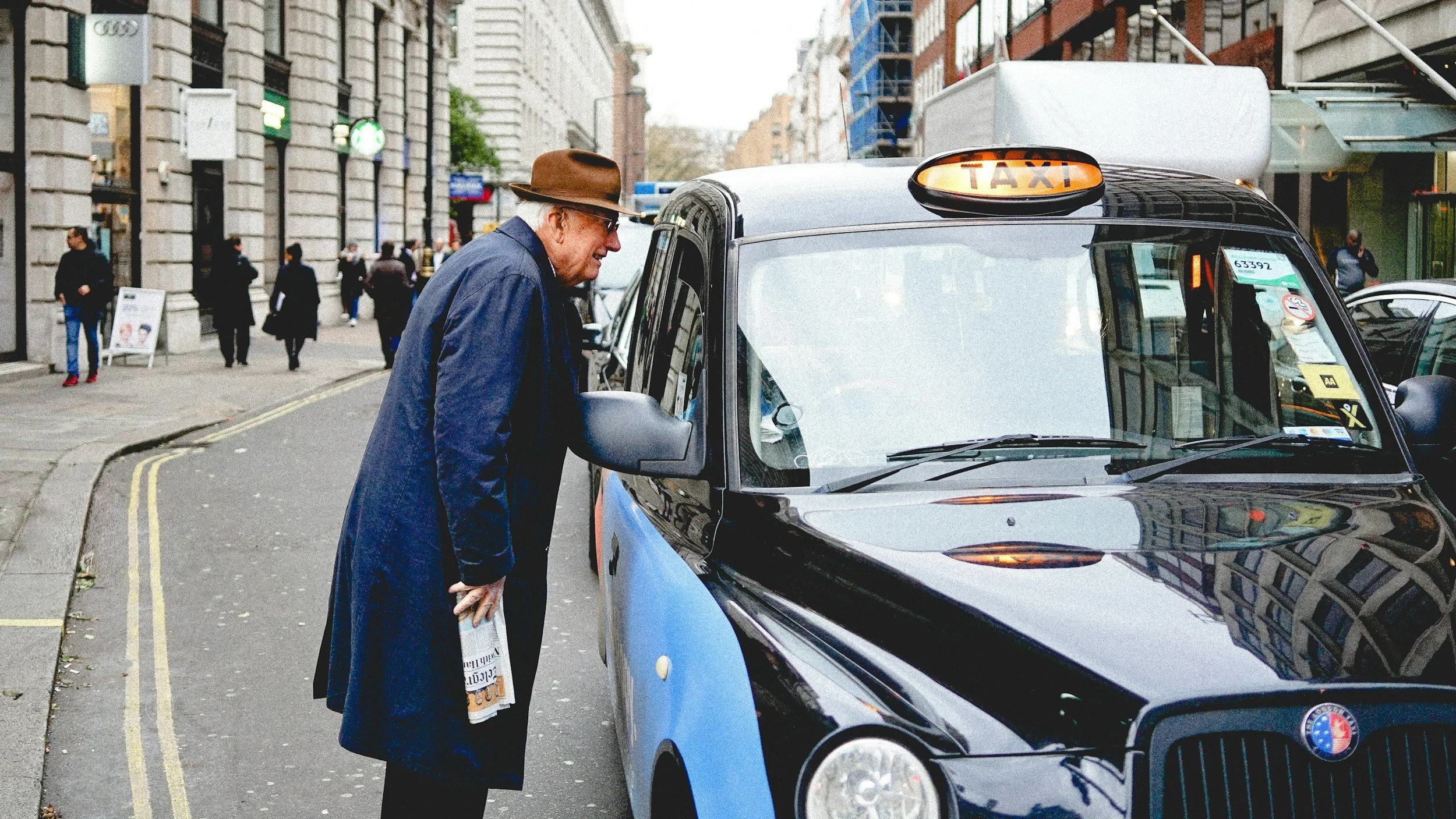 An elderly man wearing a brown hat and dark coat looks closely at a black and blue taxi parked on a city street, holding a newspaper in his hand. Several pedestrians are walking along the sidewalk in the background, with buildings and storefronts lining the street.