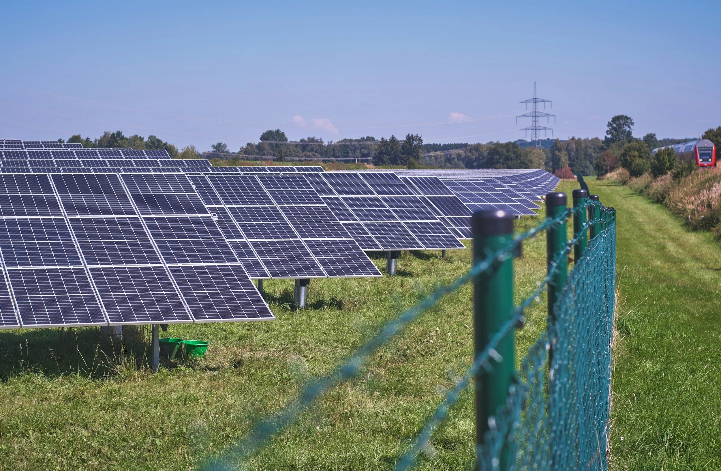 Solar panels on grassy land next to a green chain-link fence, with a train and trees in the background under a blue sky.