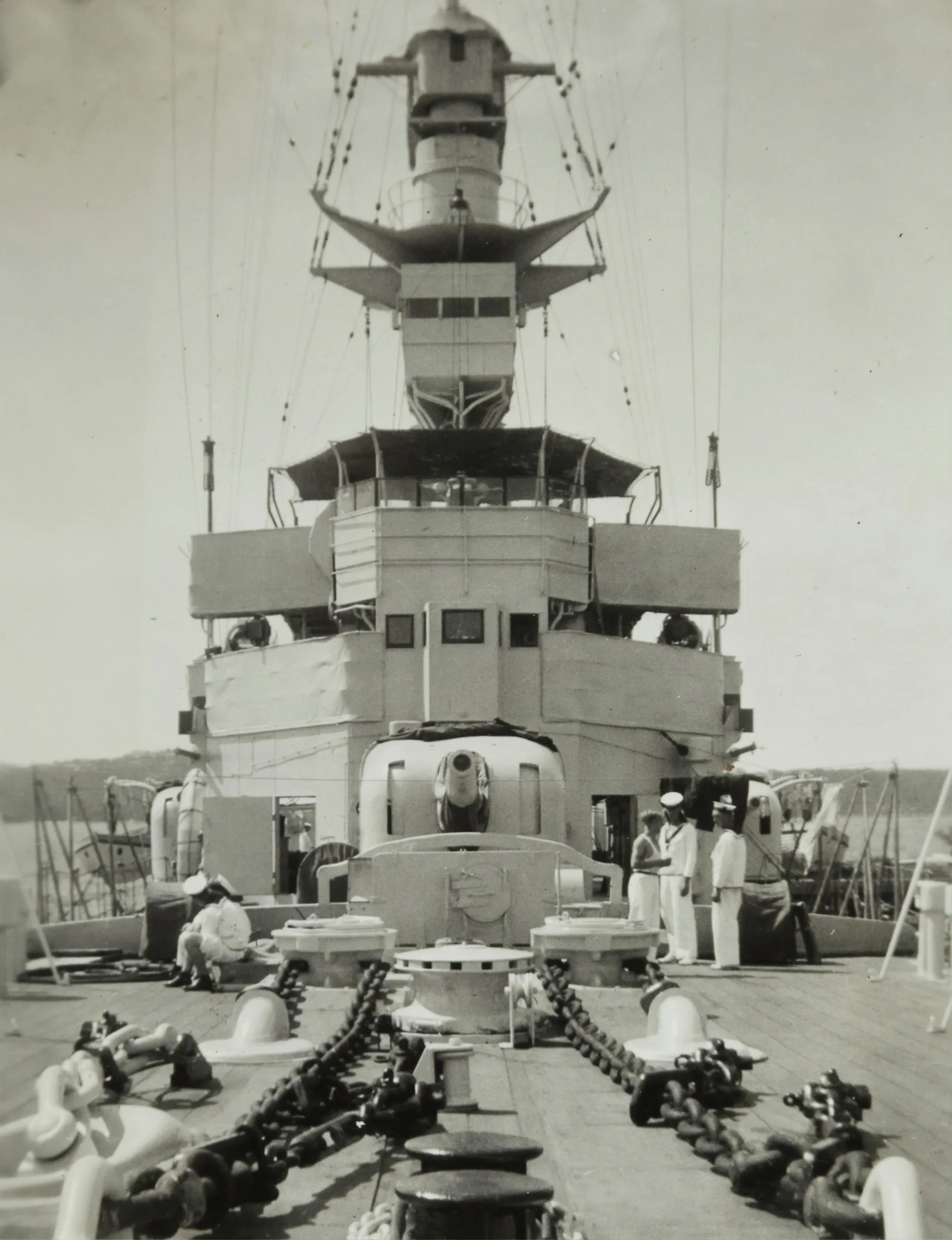 A black and white photo of a large naval battleship, focusing on its front section with crew members and ship equipment visible.