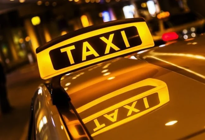 A yellow illuminated taxi sign mounted on the roof of a taxi cab at night, with its reflection visible on the car's shiny surface.