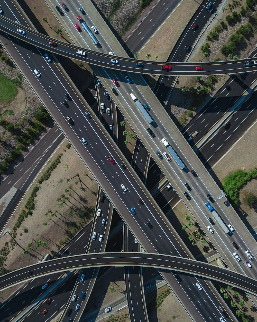 Overhead view of multiple intersecting highways with numerous cars and trucks, with shadows cast on the roads and surrounding areas with some greenery.