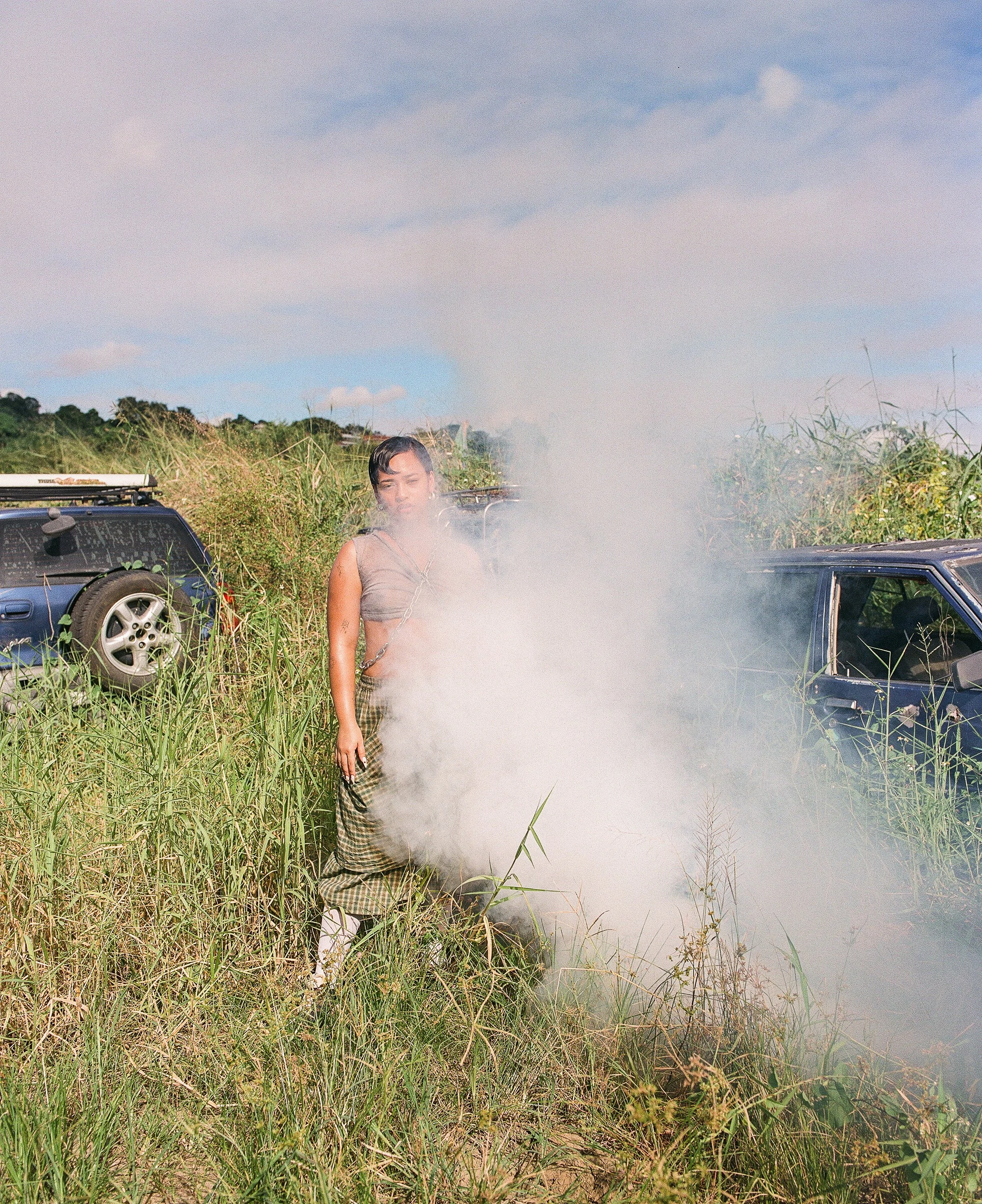 A woman standing outdoors in a grassy field with tall green grass and bushes, near two black cars, with a blue sky and some clouds in the background, and smoke or steam rising in front of her.