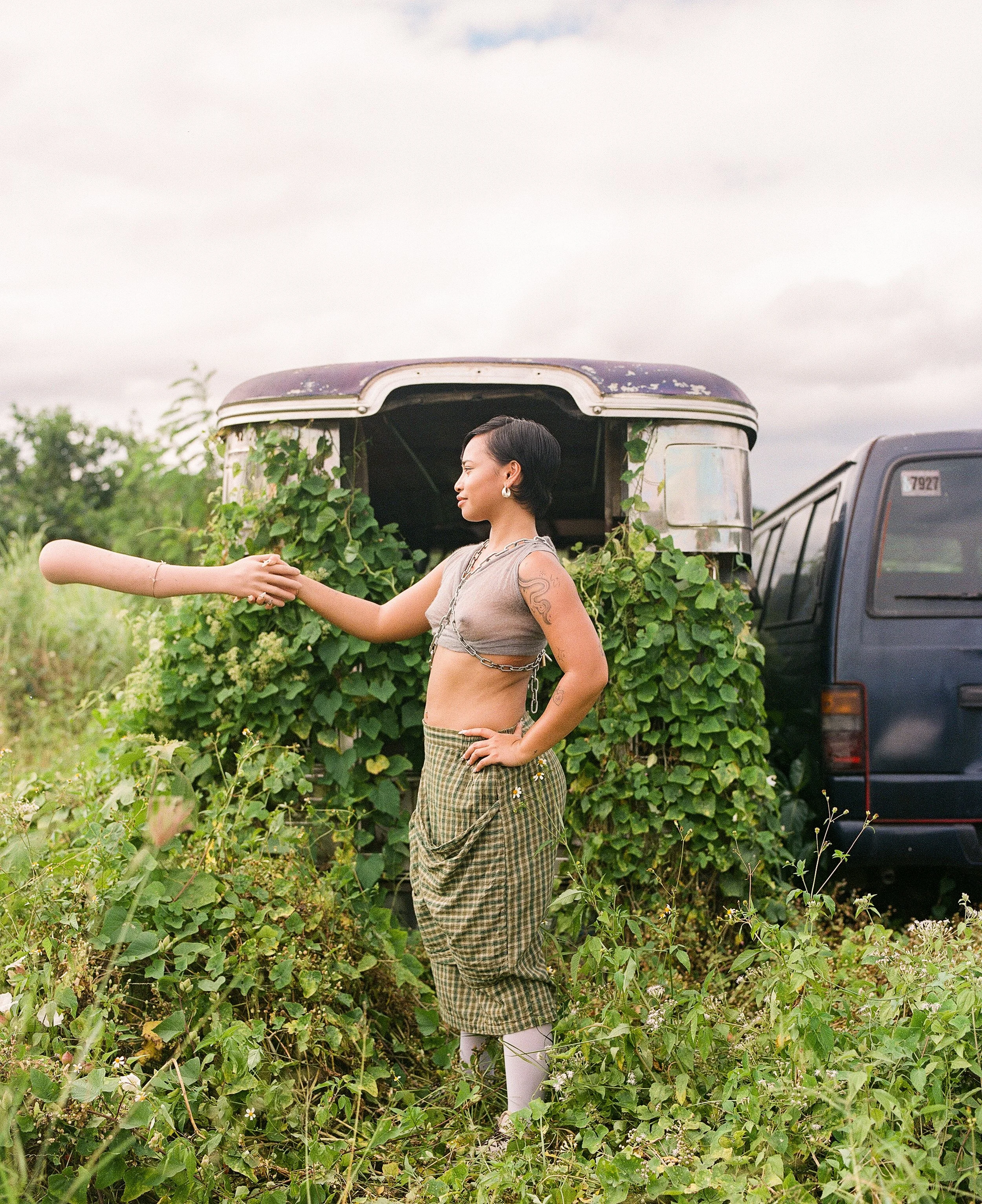 A woman with dark hair and earrings standing in a lush, green field, holding hands with another person out of the frame. Behind her is an old, weathered vehicle with an overgrown trailer filled with plants.
