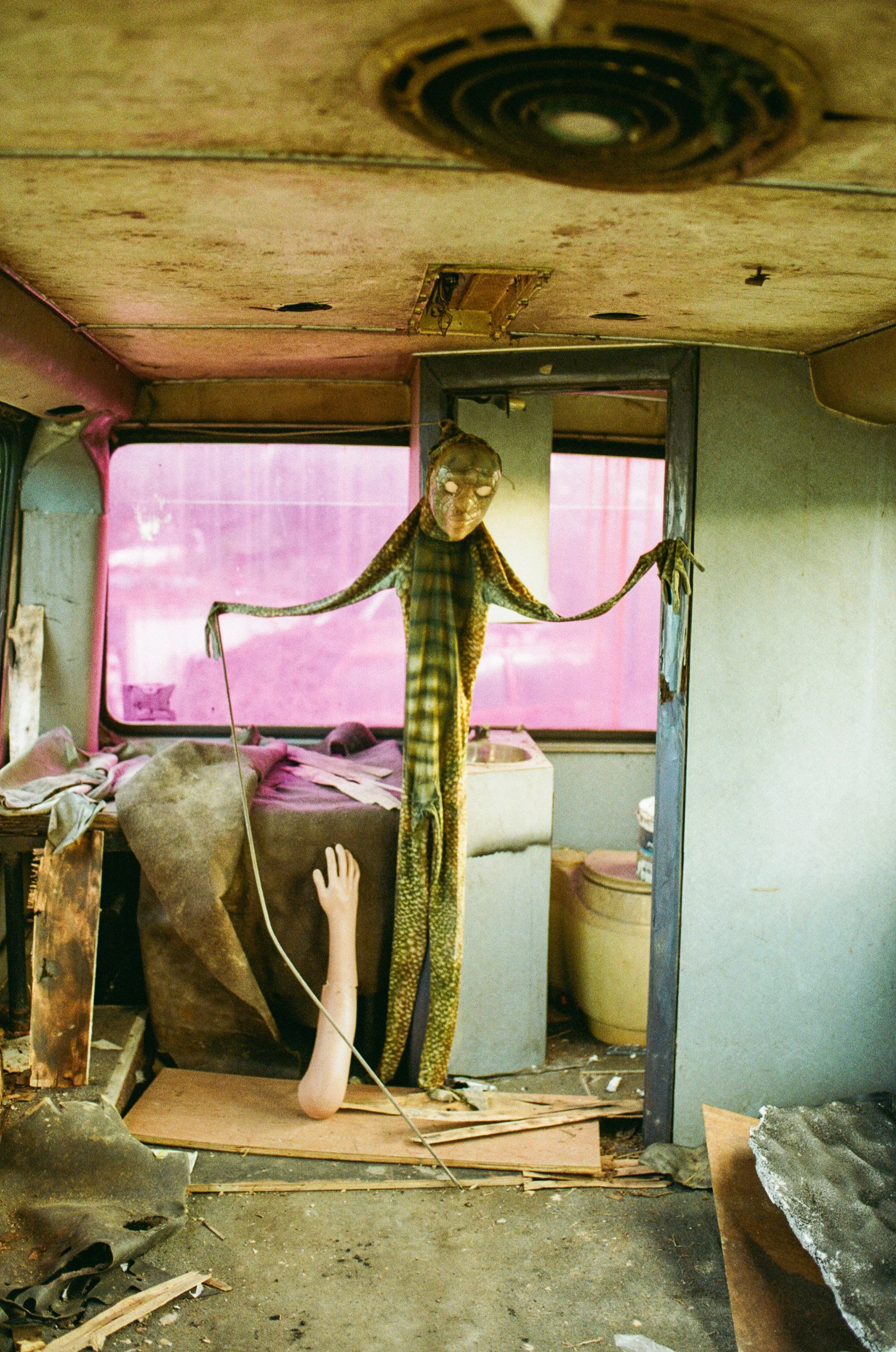 A broken and abandoned room with debris on the floor, an upside-down doll or mannequin with long limbs, and a dark, surreal atmosphere.