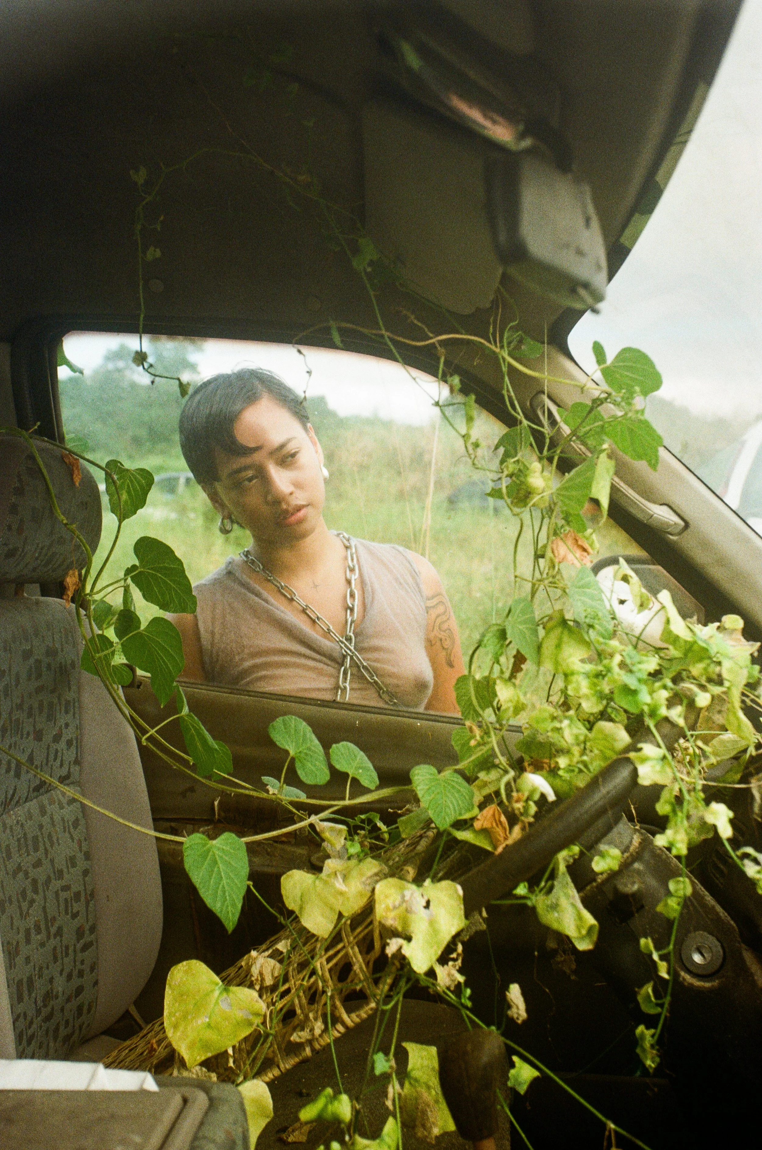 A young man with short dark hair, wearing earrings, a necklace, and a tank top, looking down inside a vehicle surrounded by green vines and leaves.