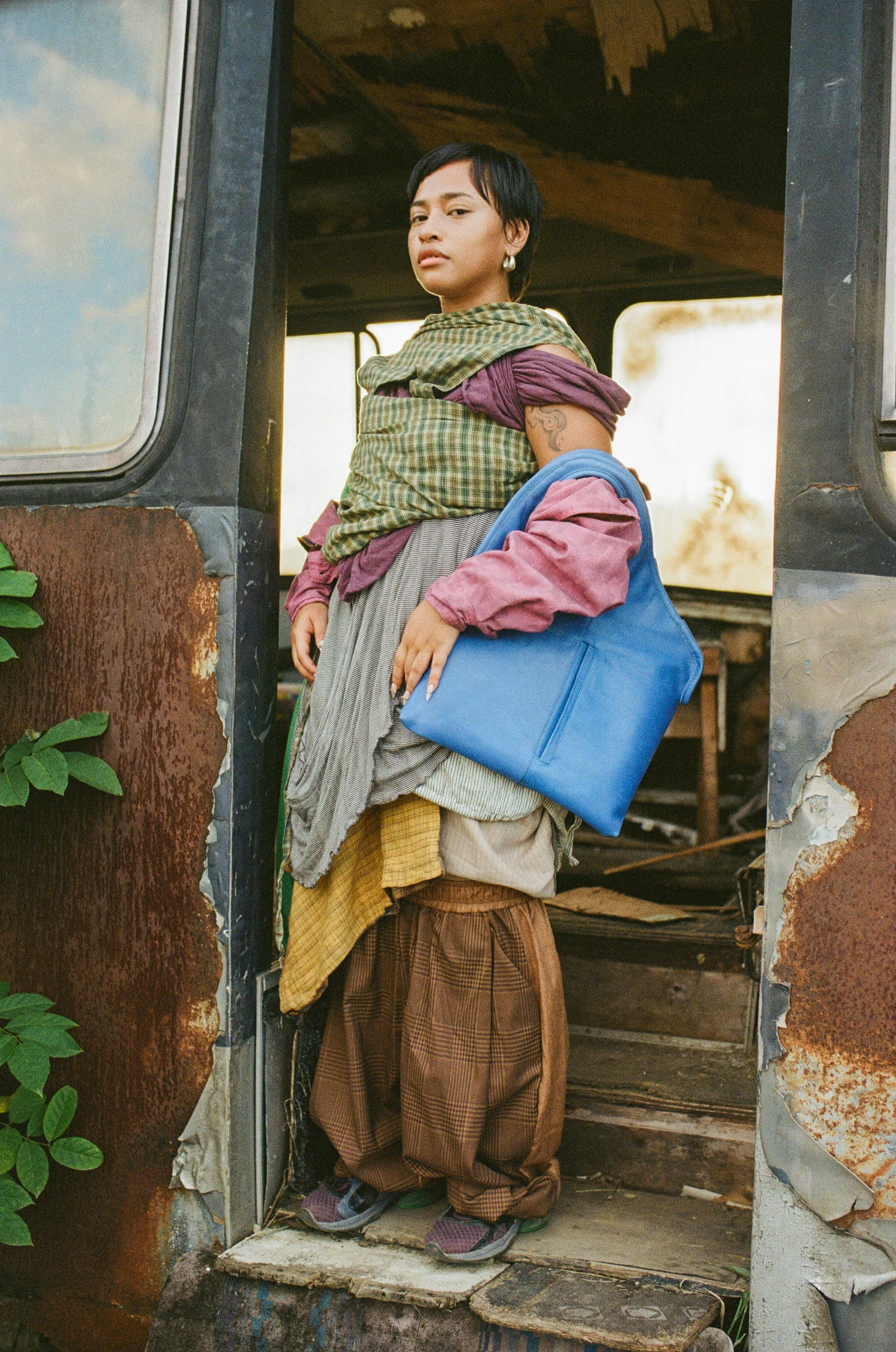 A young woman standing at a door frame with her hands resting on her hips, looking confidently at the camera. She is wearing layered clothing that appears comfortable and eclectic, including a scarf, earrings, and patterned pants. The background shows an interior space with unfinished, rustic walls and natural lighting.