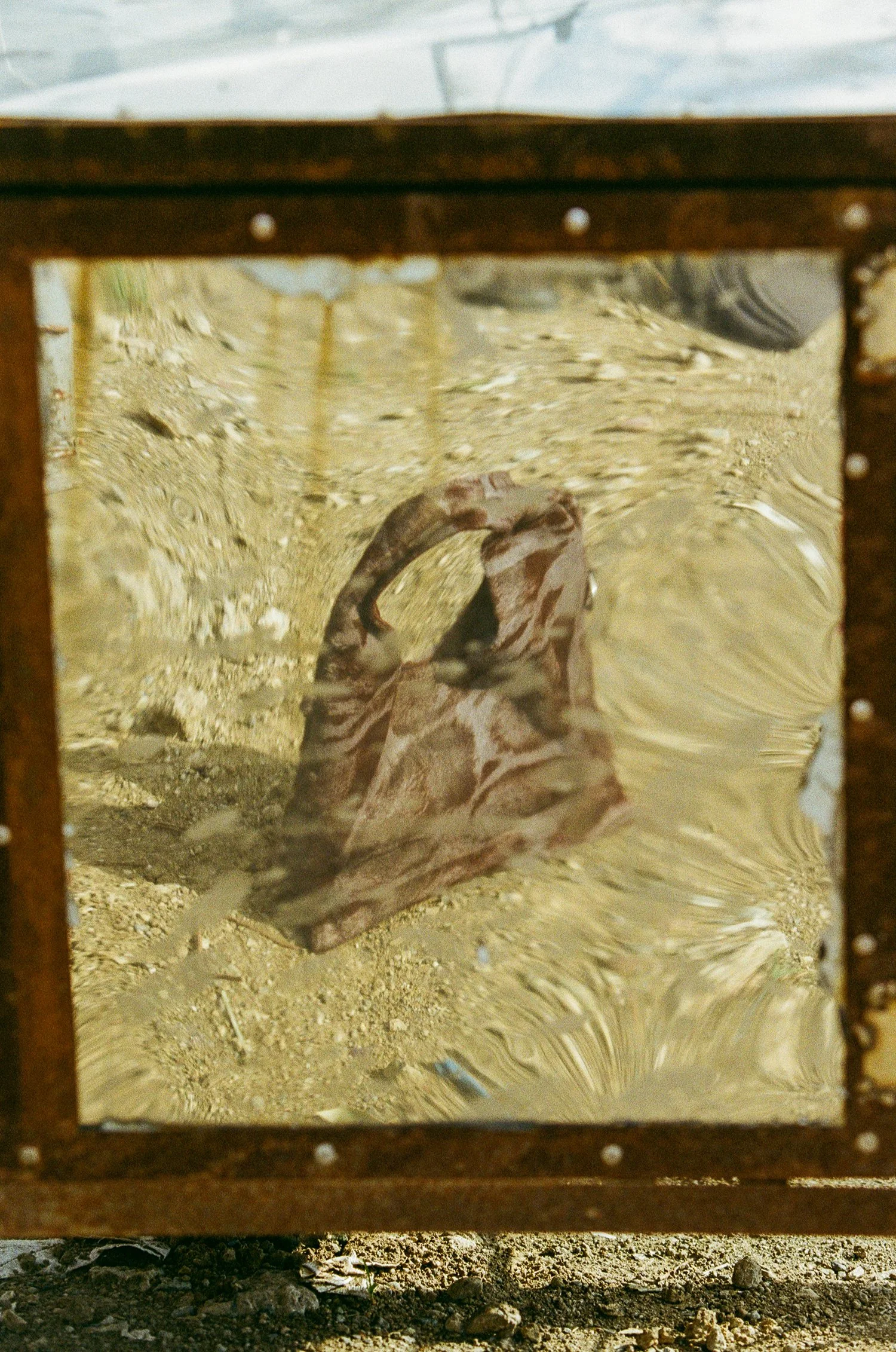 A giraffe lying down on a sandy ground viewed through a glass panel framed by a metal structure.