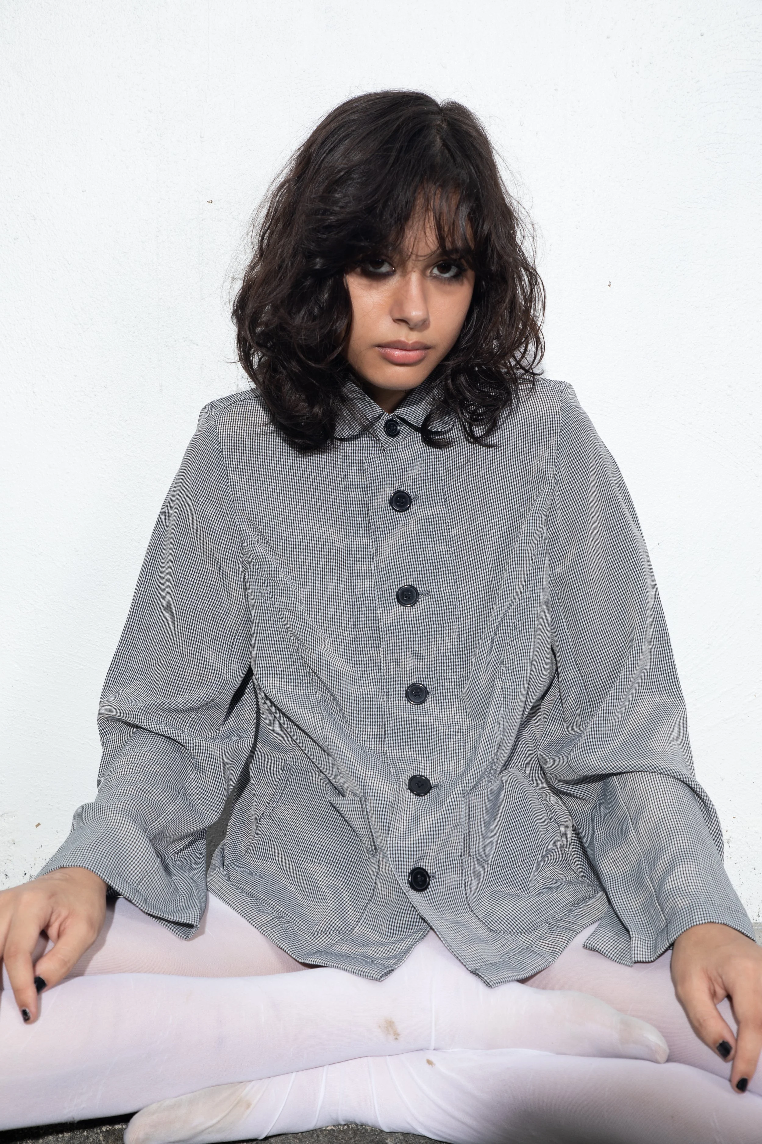A woman with dark curly hair and dark eye makeup sitting on a white couch against a plain white wall, wearing a grey checkered shirt and white leggings.