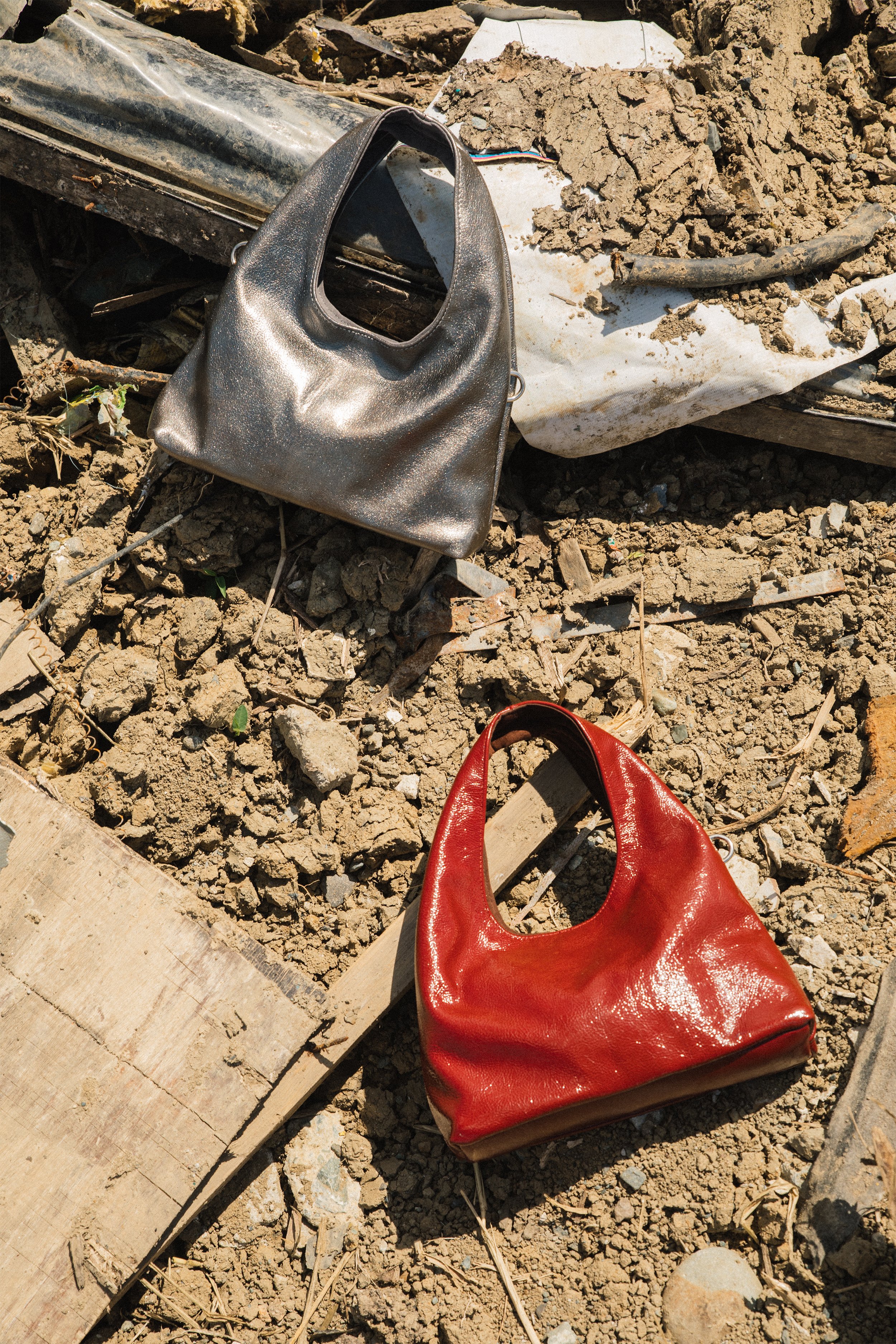 Red patent leather purse and gray handbag placed on dirt and rubble with wooden planks, metal debris, and a white tarp in the background.