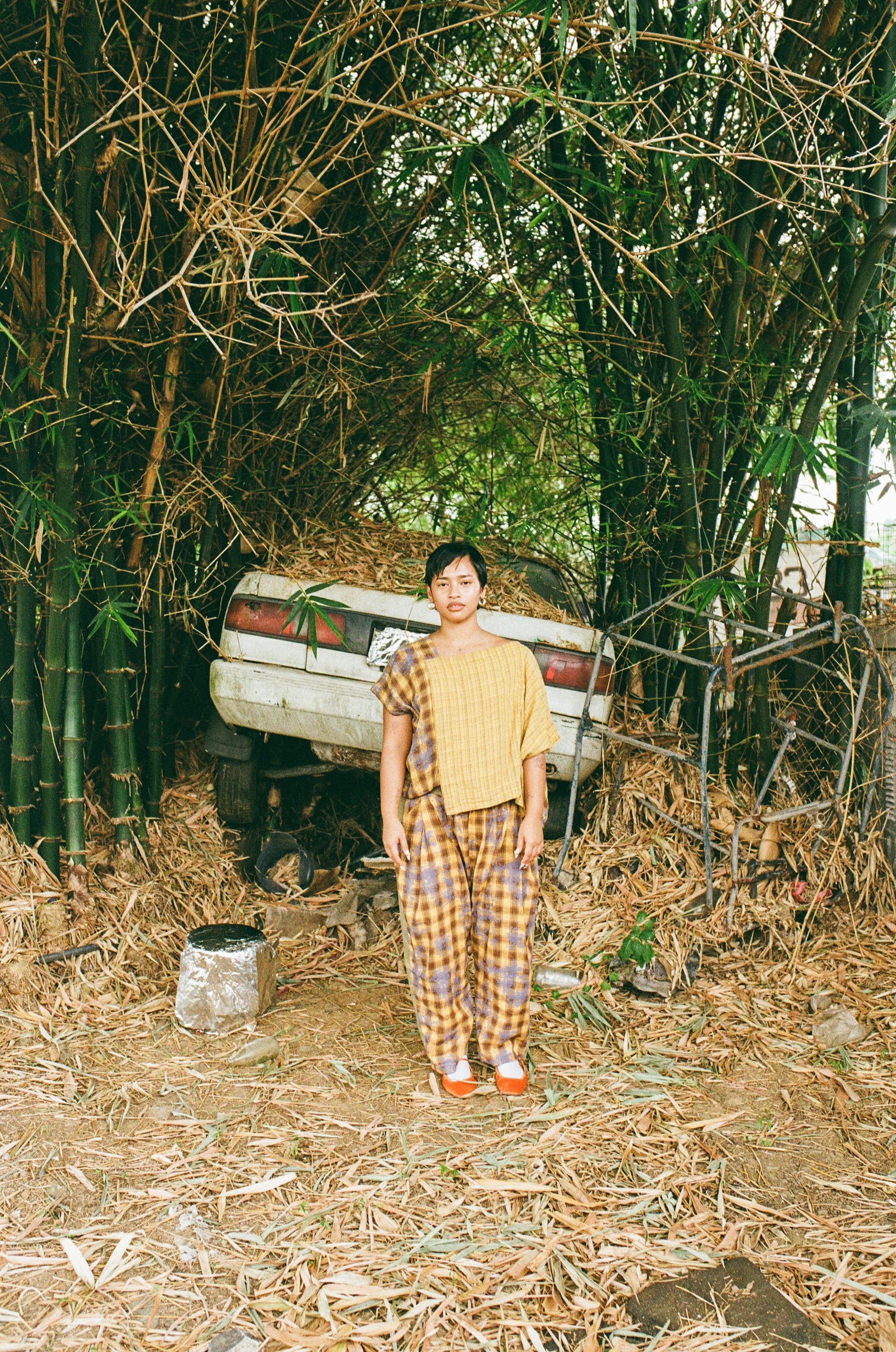 A young woman in plaid clothes and white shoes standing in front of a crashed, overgrown car surrounded by tall bamboo and dry leaves.
