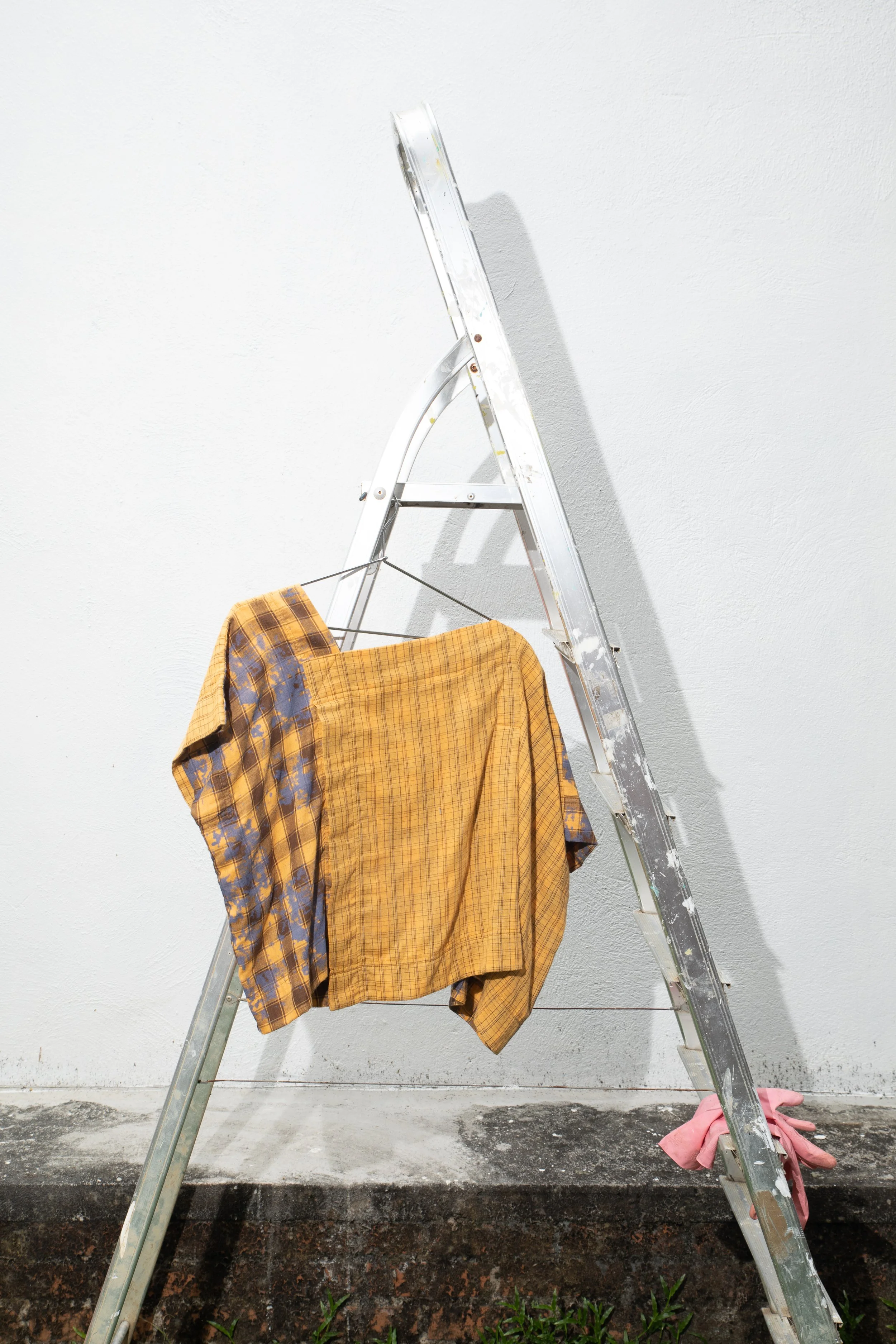 A ladder leaning against a plain white wall with a yellow and purple plaid shirt hanging on it. A pink cloth is at the base of the ladder on the ground.