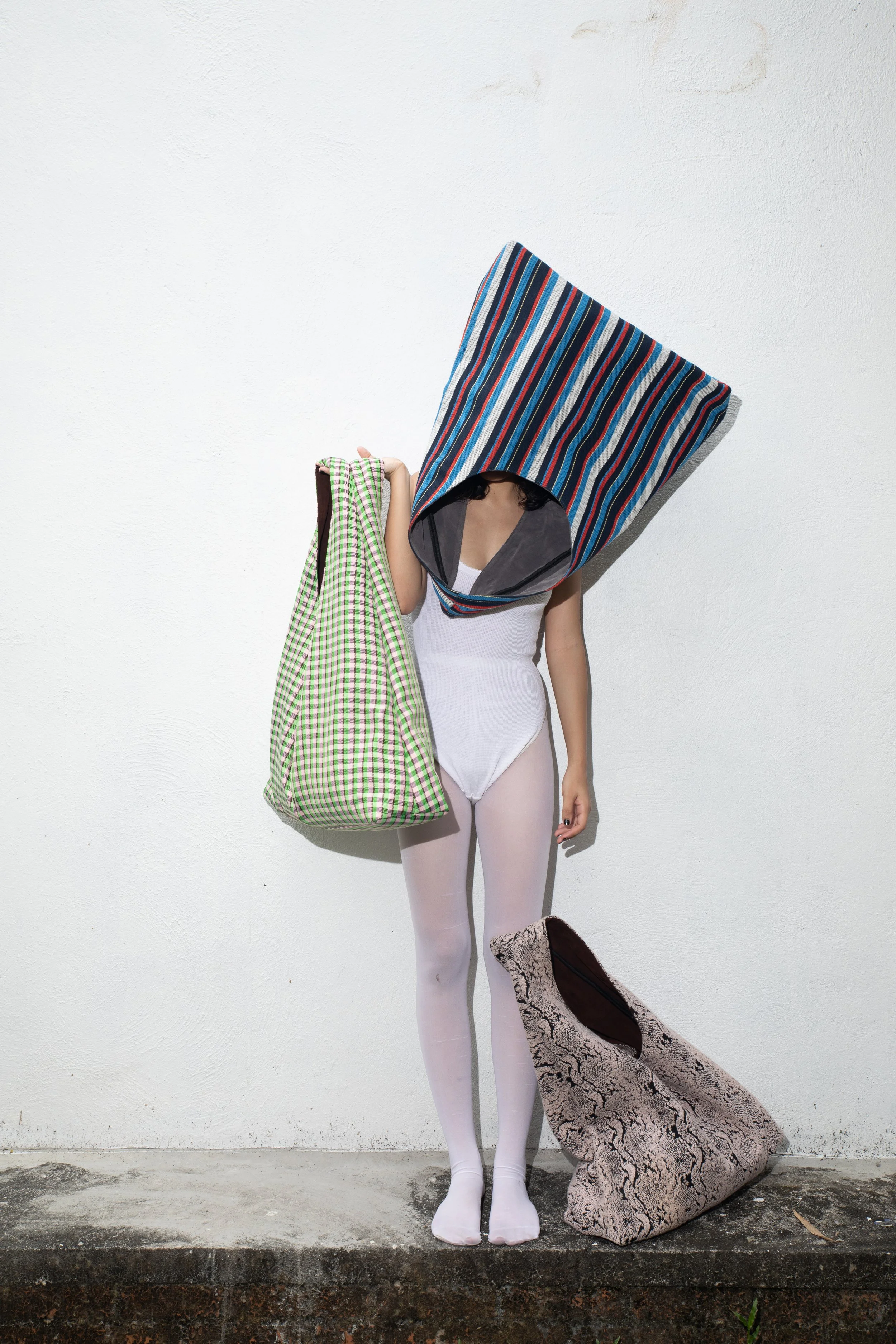 Person with a large striped bag over their head, holding a green checkered bag, standing against a white wall with a large rock on the ground.