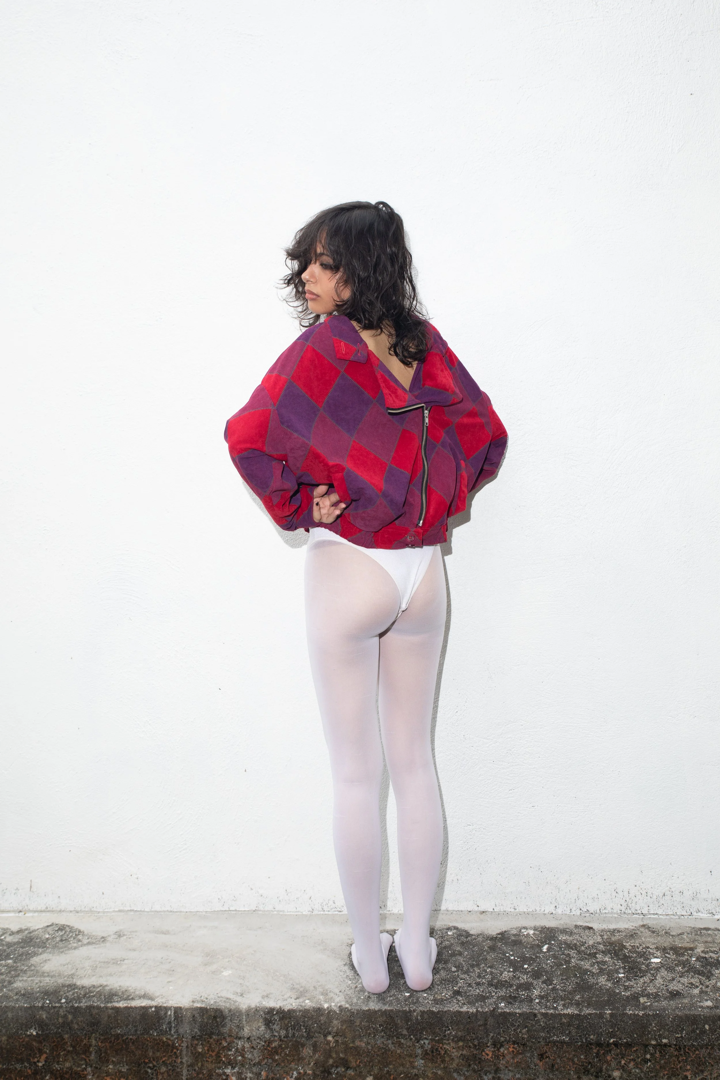 A woman with dark, curly hair wearing a red and purple checkered jacket and white tights, standing against a plain white wall, facing away with her head turned to the side.