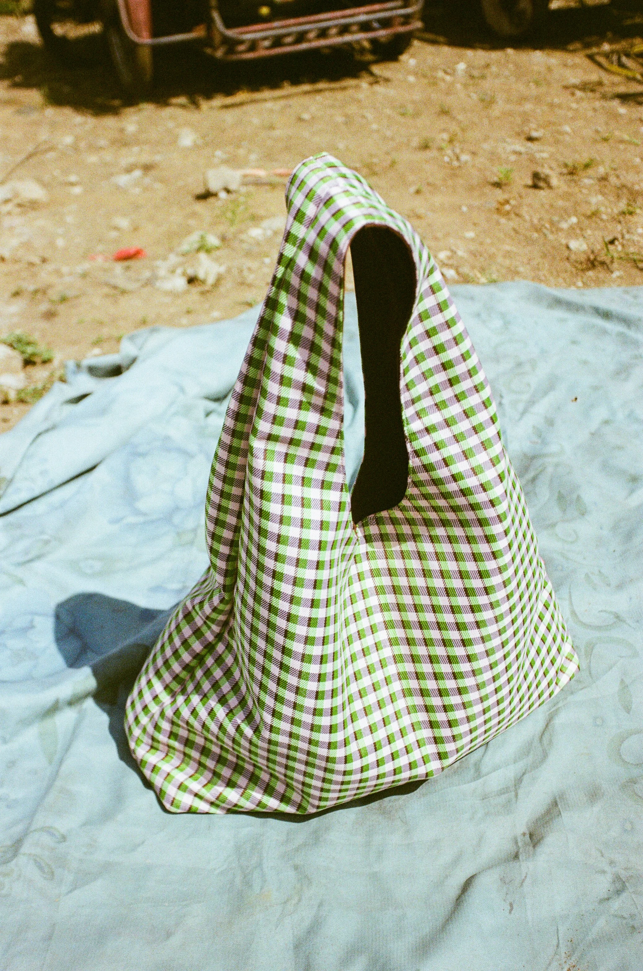 A person wearing a green and white checkered garment standing outdoors on dirt ground, with a cloth or sheet on the ground and some objects blurred in the background.