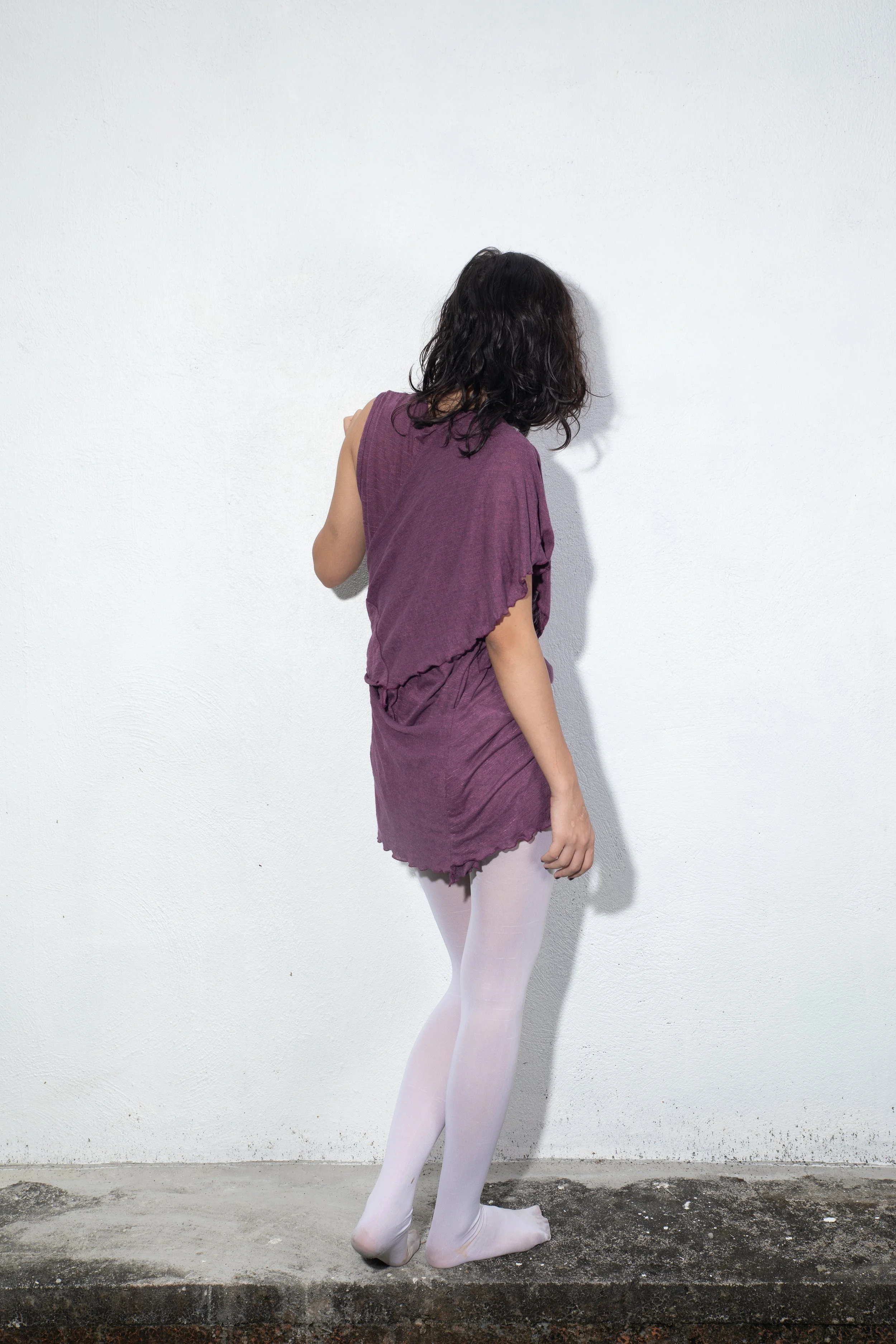 A person with curly dark hair wearing a purple dress and white tights, standing against a plain white wall, with only the head, arms, and legs visible.