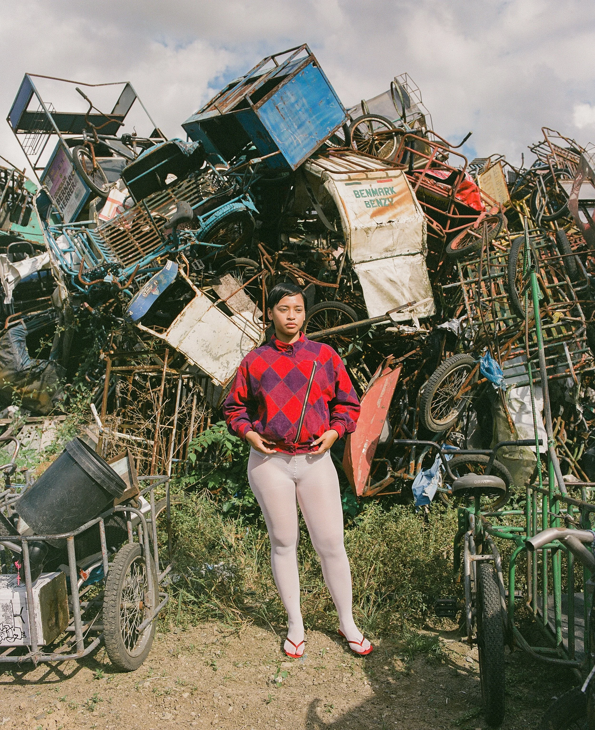 A woman stands outdoors in front of a large pile of rusty bicycles and carts. She is wearing a red and purple checkered jacket, white pants, and red sandals. The sky is partly cloudy with patches of blue visible.
