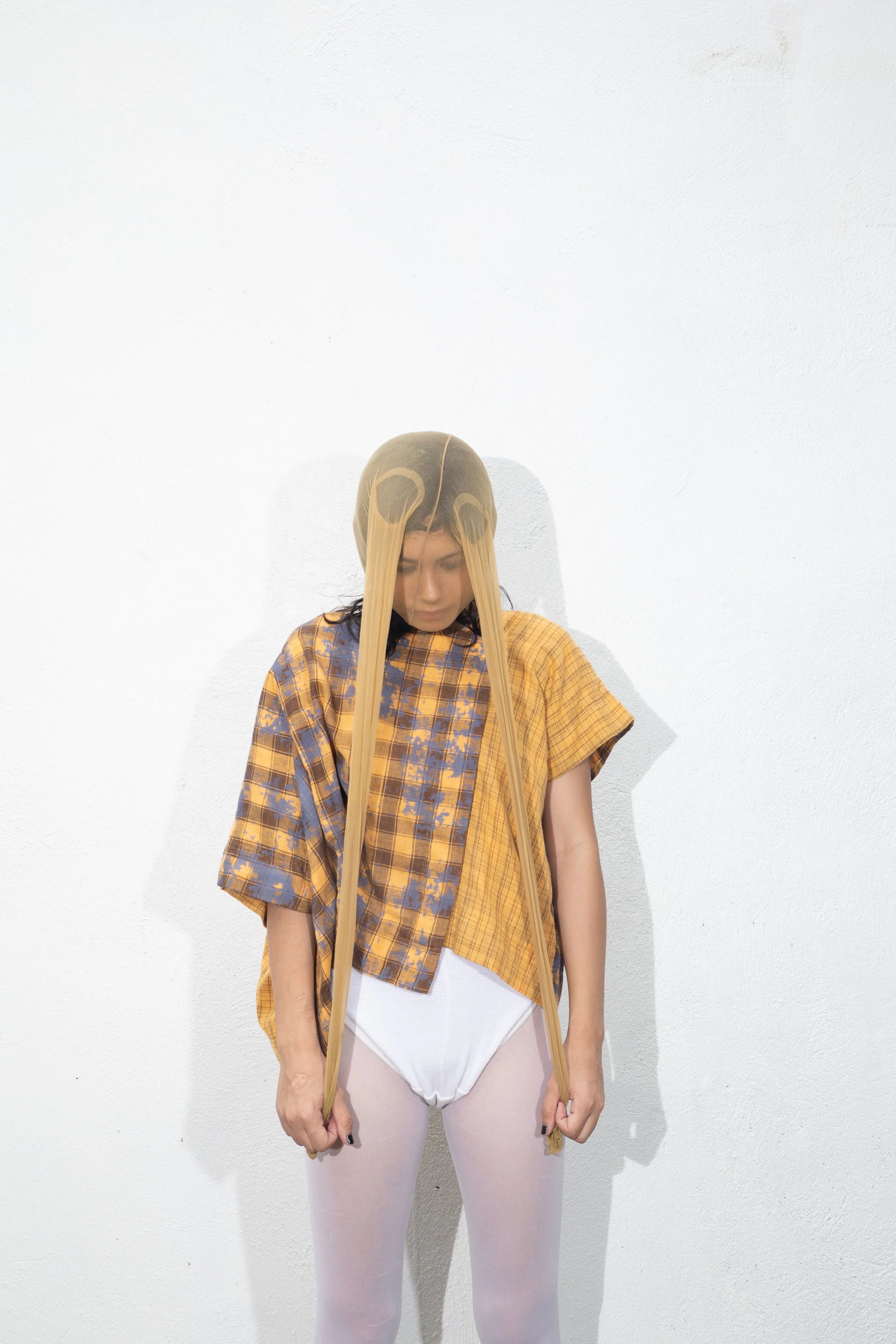 A woman standing against a white wall, wearing a gold and blue plaid shirt, white underwear, and pale pink tights, with a wet hairdo softened by a beige head covering resembling a skull.