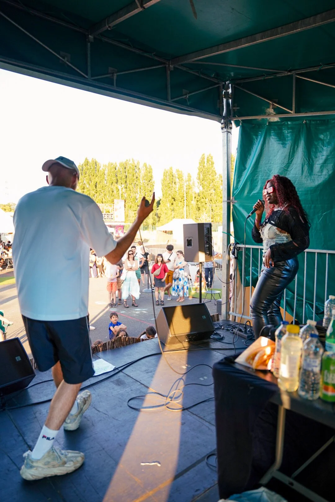 Une scène de concert en plein air avec deux performeurs sur scène, une femme chantant avec un microphone et un homme en mouvement, face à un public diverse.