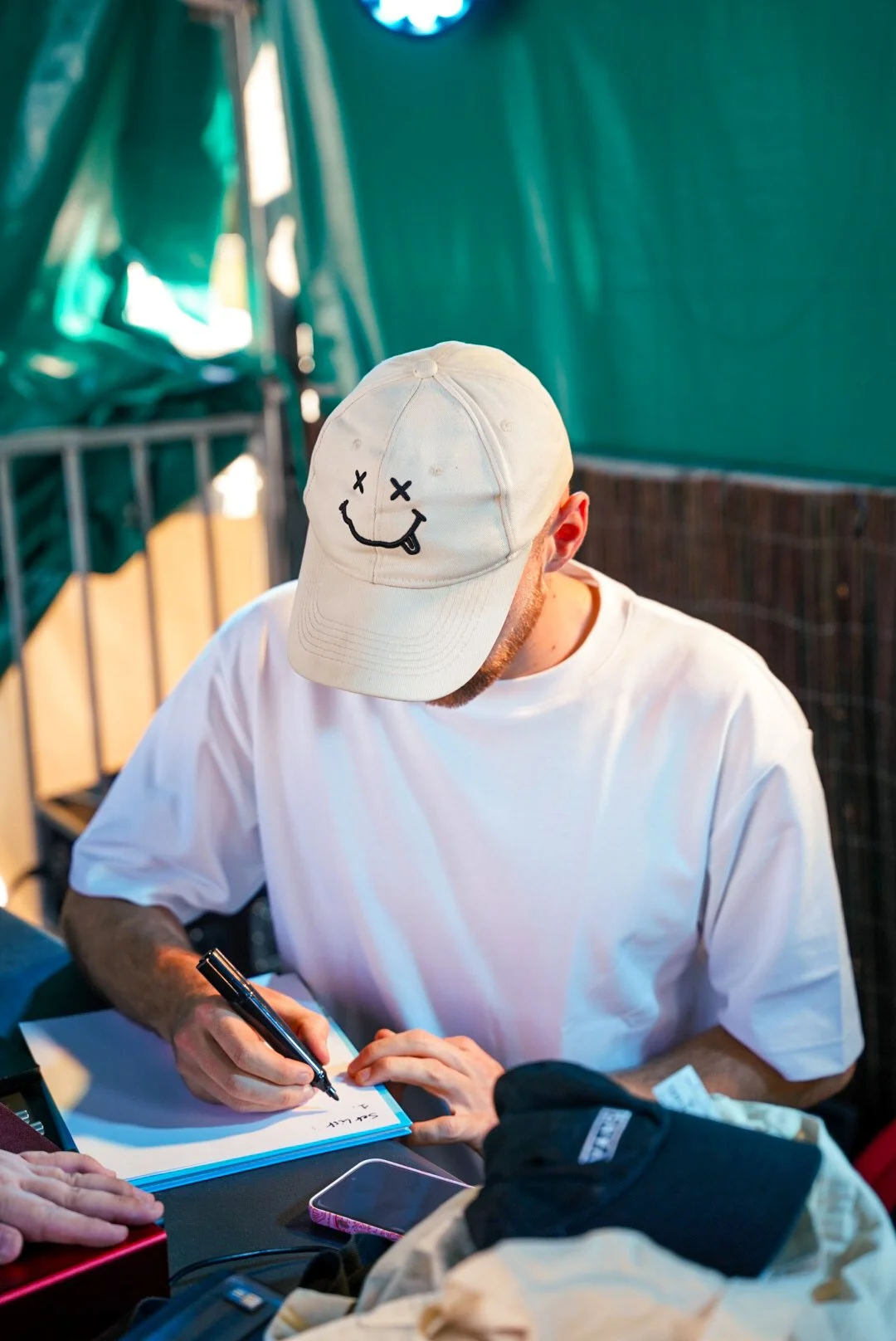 Un homme portant une casquette blanche avec un visage dessiné et un T-shirt blanc, écrit dans une feuille, dans un environnement extérieur.
