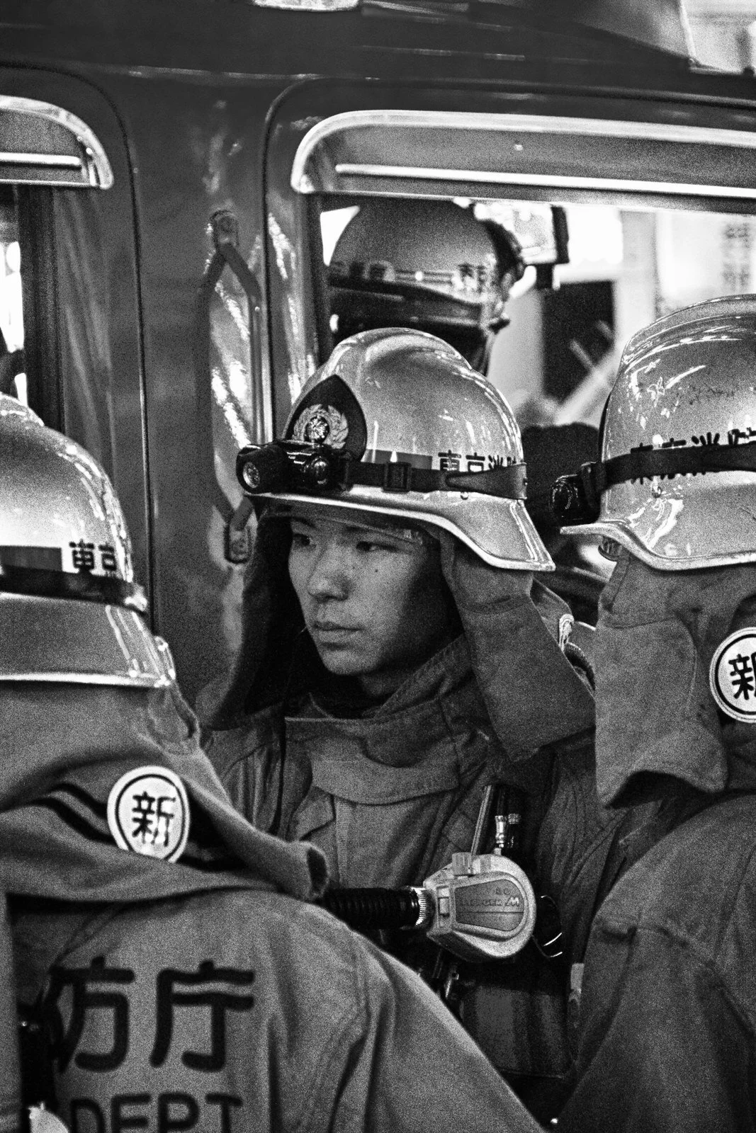 A female firefighter adjusting her helmet, surrounded by other firefighters, with a fire truck in the background, in black and white.