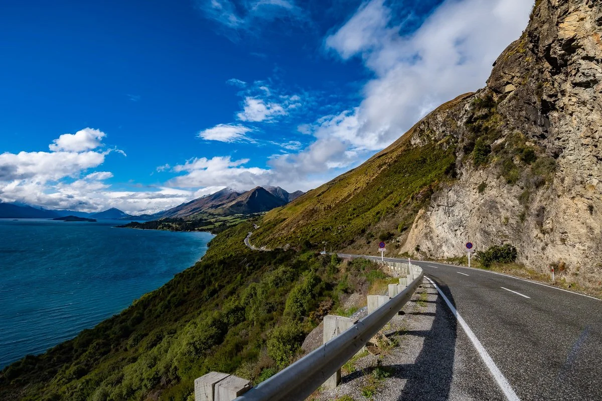 A winding coastal road runs along a mountainside overlooking a lake, with mountains in the distance under a partly cloudy blue sky.