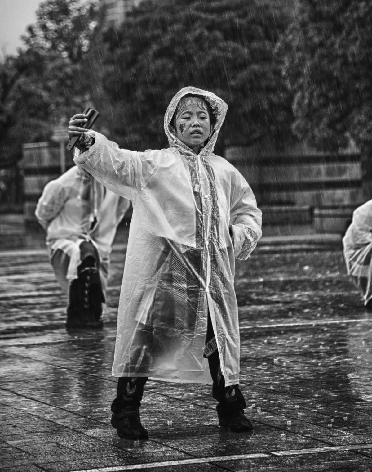 A young person wearing a raincoat and boots, standing in the rain with a serious expression, holding a cellphone in one hand, with other people also dressed for rain in the background.