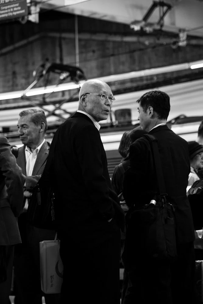 Black and white photo of people waiting on a train platform, engaged in conversation, with a train and station infrastructure in the background.
