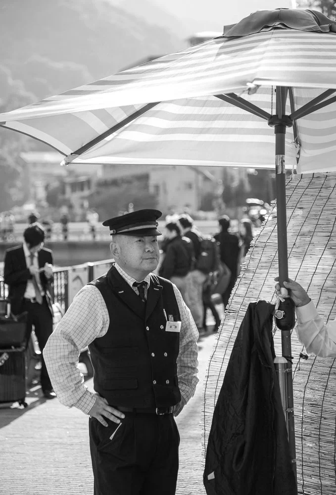 A man dressed as a security guard stands outdoors under a large striped umbrella, with others walking in the background.