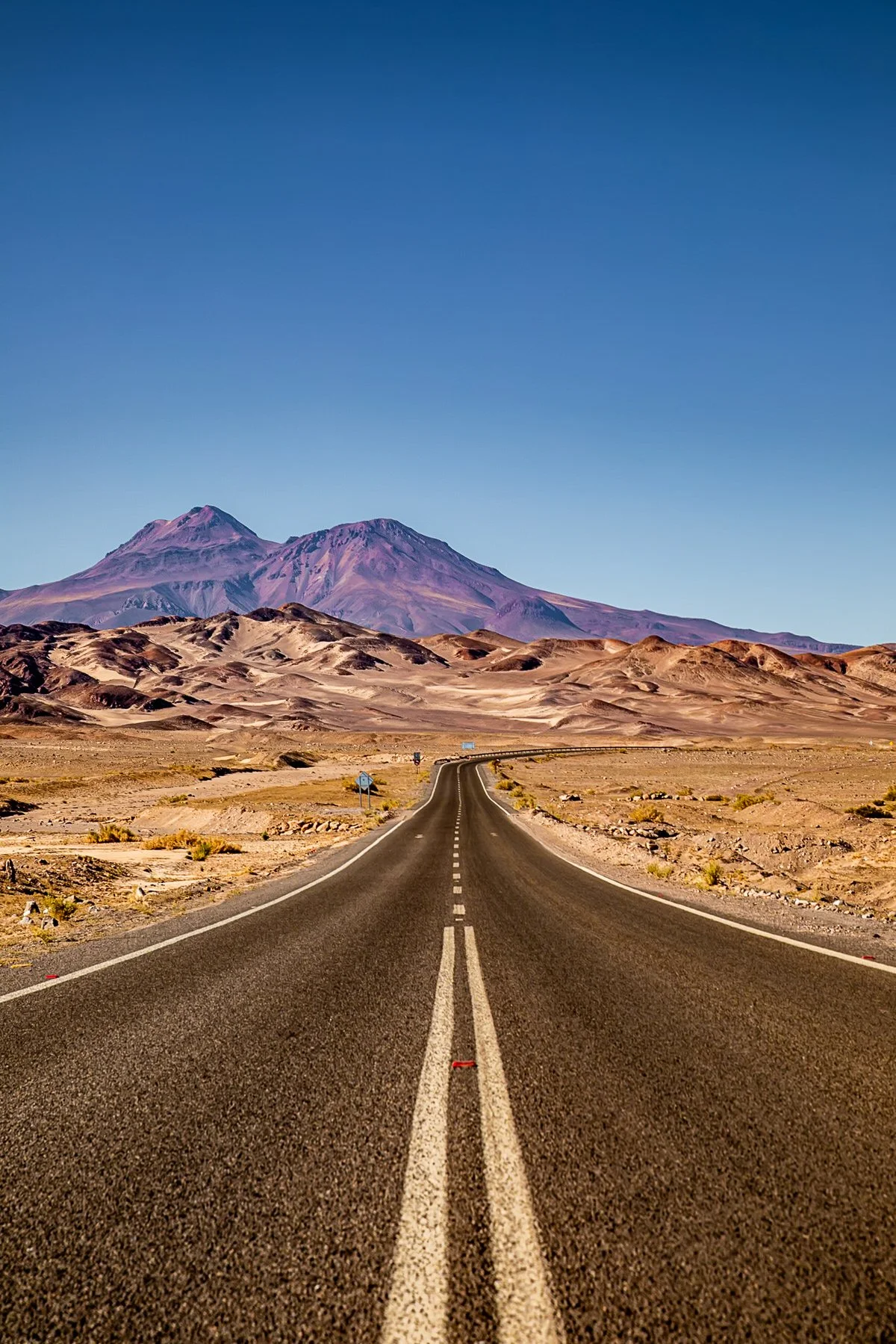 A deserted straight asphalt road cutting through a desert landscape with mountains in the background and a clear blue sky.
