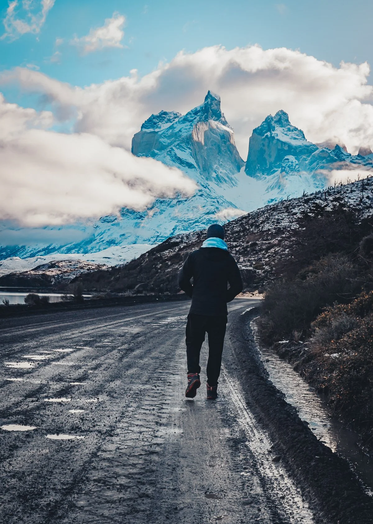 A person dressed in winter clothing, walking on a dirt road in a mountainous area with snow-covered peaks and cloudy sky in the background.