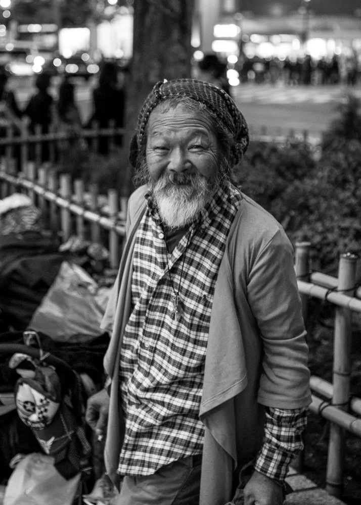Black and white photo of an elderly man with a beard and braided hair, smiling outdoors in a city park with people and city lights in the background.
