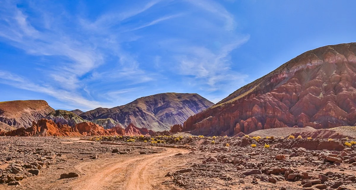 Desert landscape with colorful hills and a dirt road under a blue sky with wispy clouds.