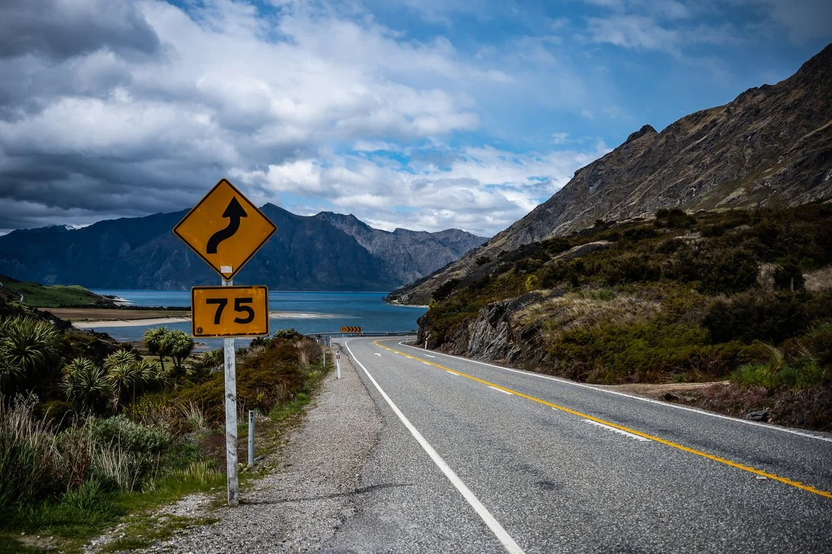 A winding mountain road with a yellow curve warning sign and a 75 km/h speed limit sign, with lakes and mountains in the background.