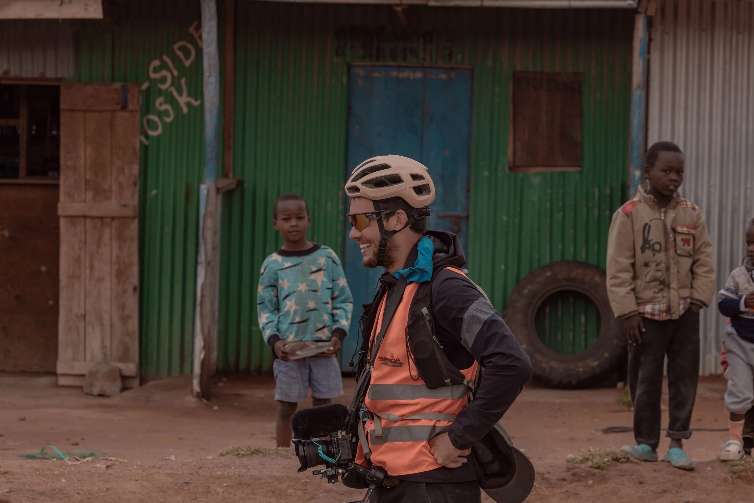A man wearing a helmet, sunglasses, and an orange safety vest smiling and talking to children in front of a green and rusty corrugated metal building.