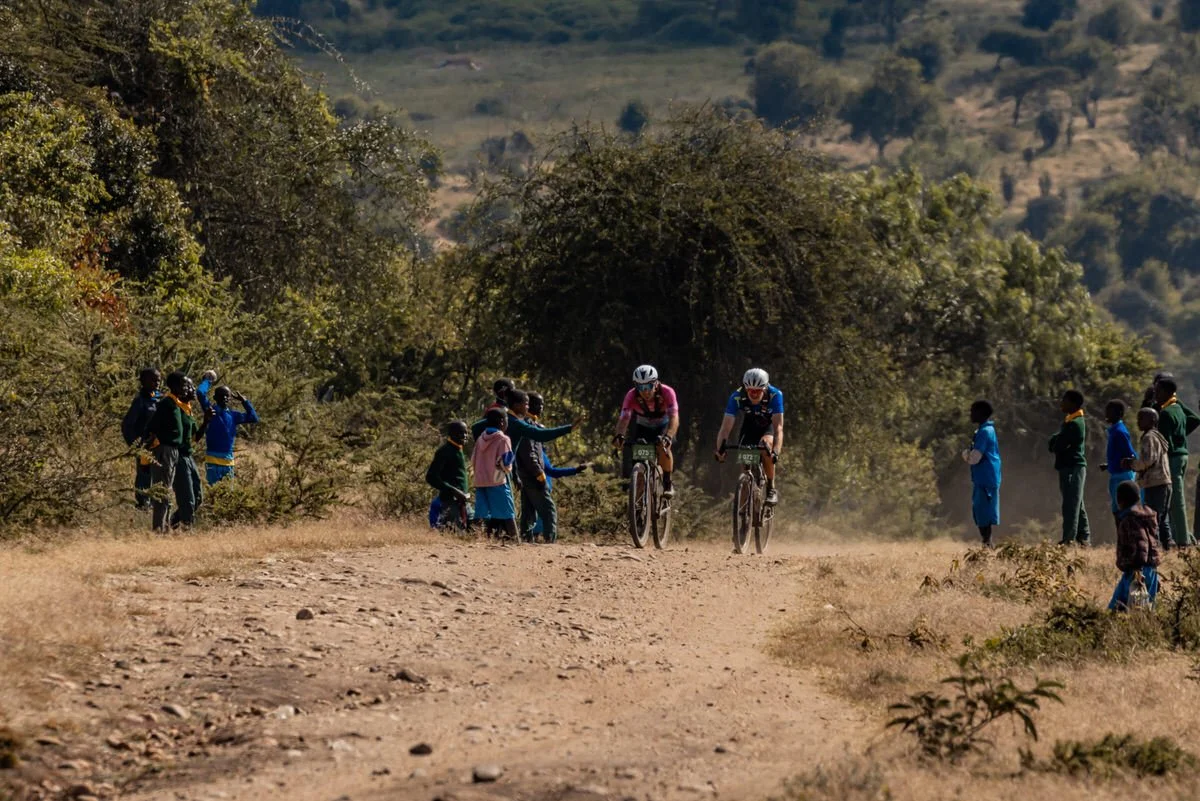 Two cyclists racing on a dirt trail surrounded by spectators in a rural setting with hills and trees in the background.