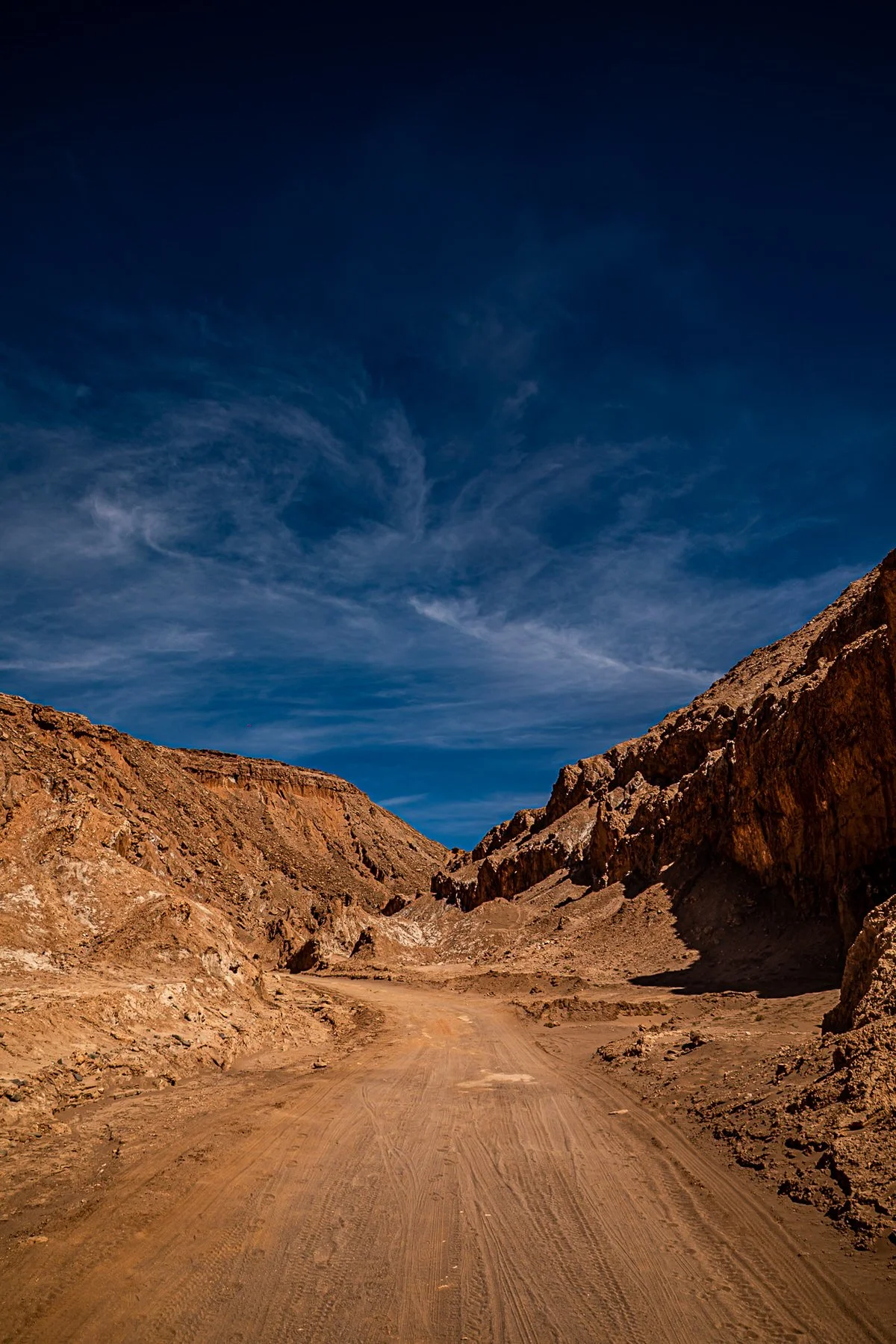 A dirt road winding between rocky, sandy mountains under a blue sky with wispy clouds.