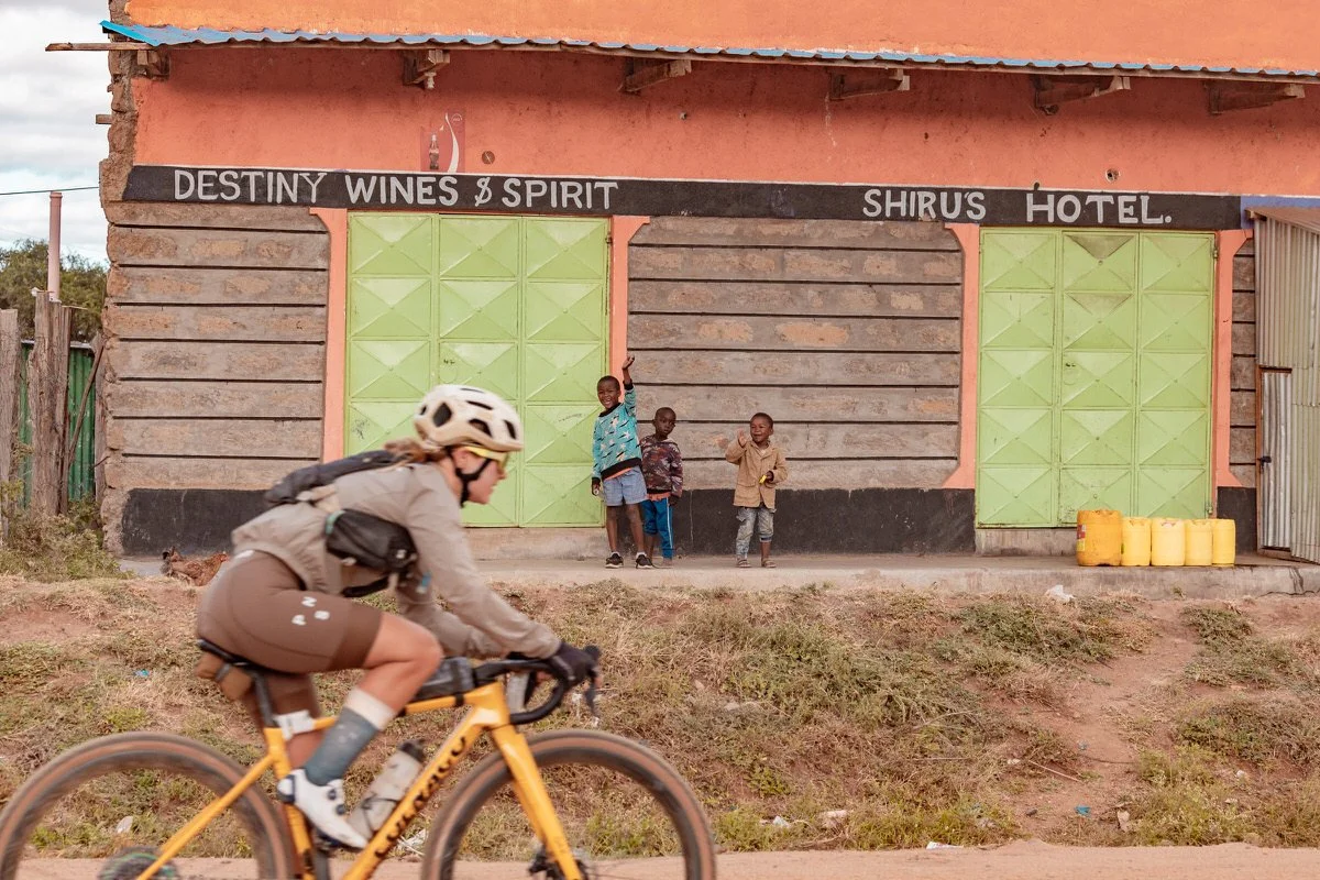 A person riding a yellow bicycle past a building with four children standing in front of painted green doors, with signage reading 'Destiny Wines & Spirit' and 'Shiru's Hotel.'