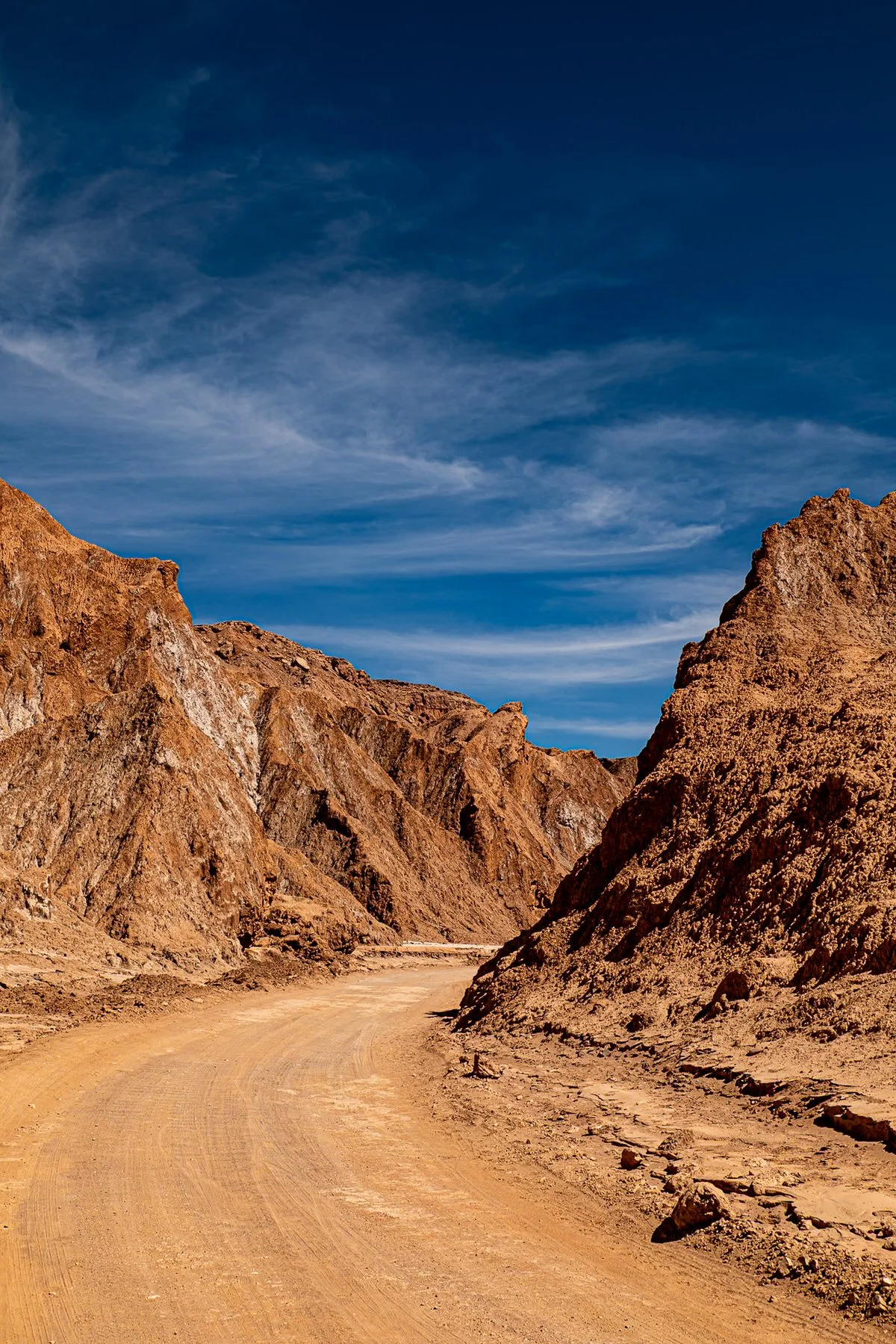 Dusty unpaved road winding through a desert canyon with rugged brown cliffs under a blue sky with wispy clouds.
