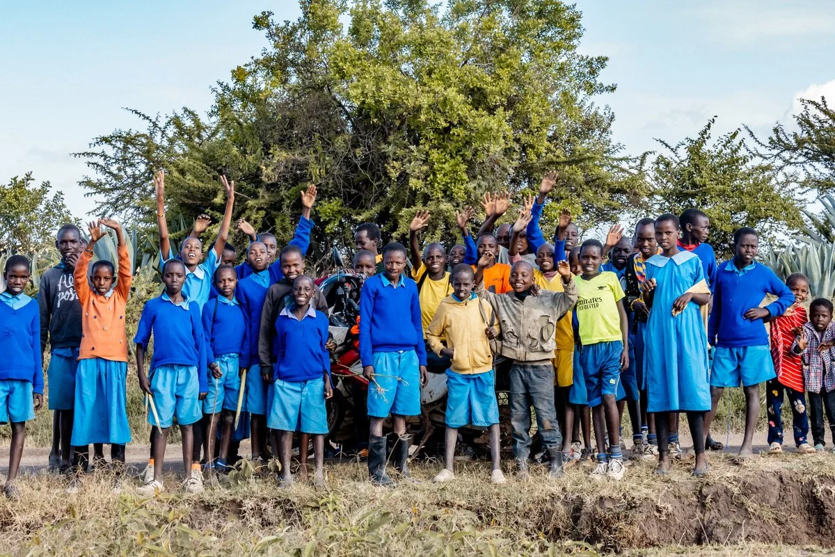 Group of children standing outdoors in front of a large bush, some raising their hands, with a motorcycle partly visible behind them.