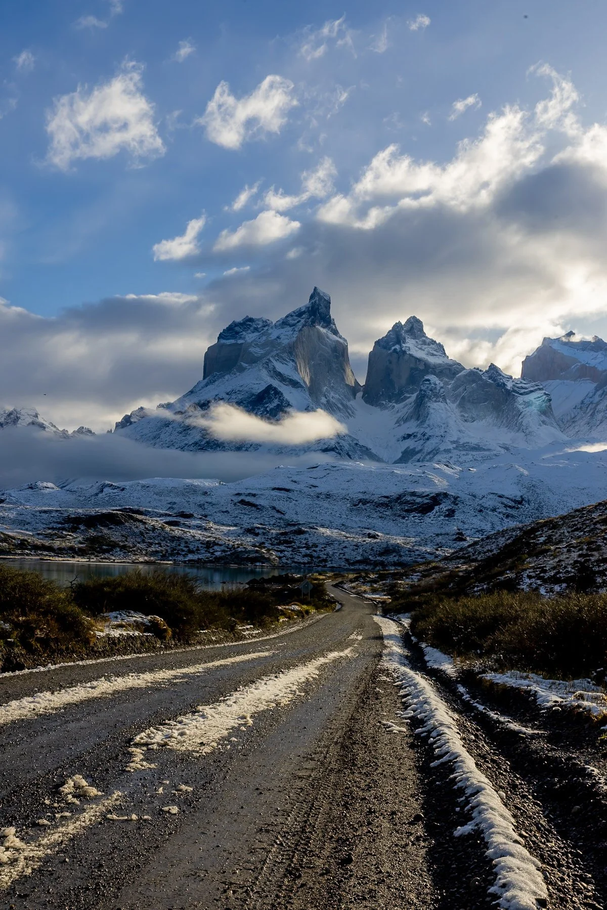 A rugged dirt road with snow patches leading towards snow-capped mountains under a partly cloudy sky.