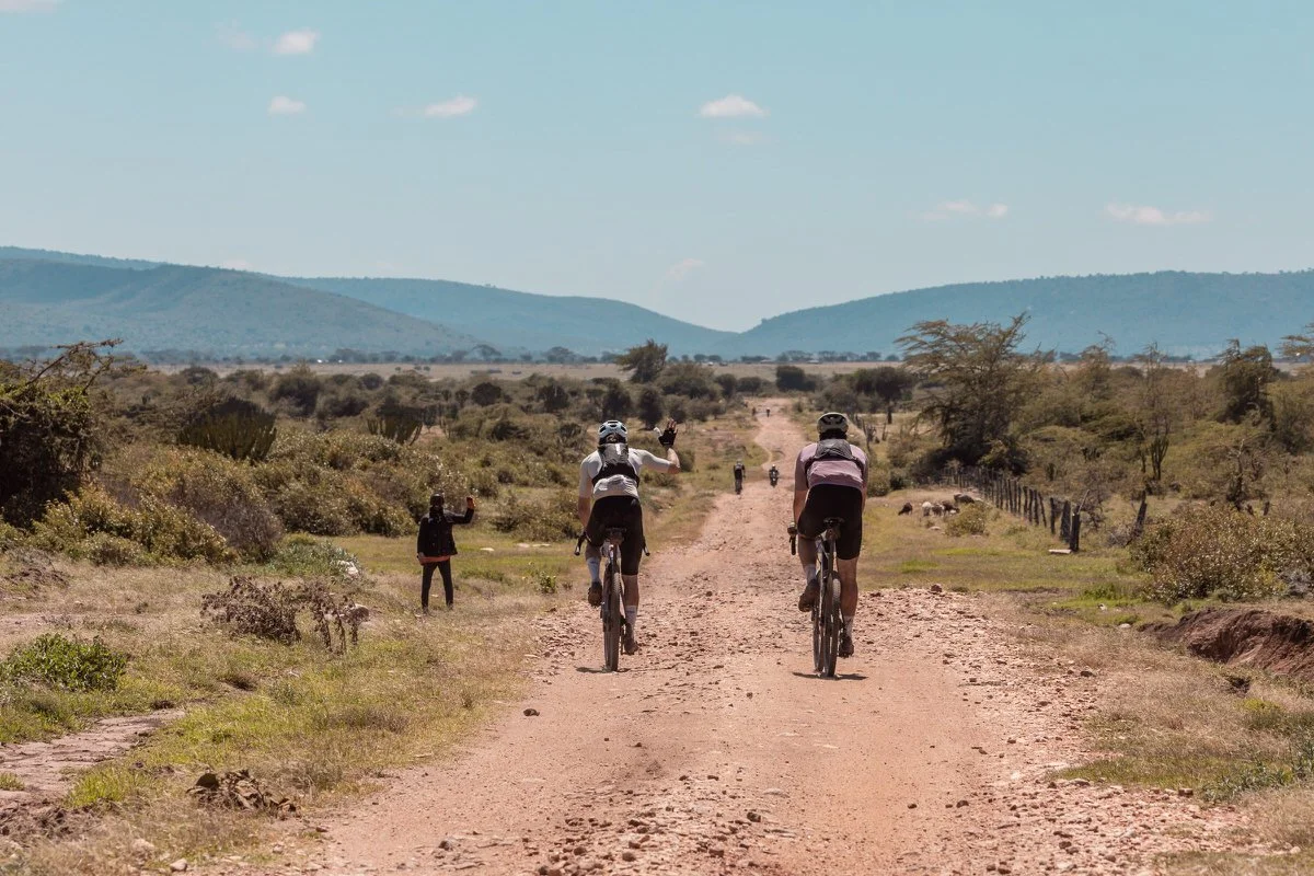 Two people riding bikes on a dirt road in a semi-arid landscape with distant hills, some people standing along the roadside, and a bright blue sky with a few clouds.