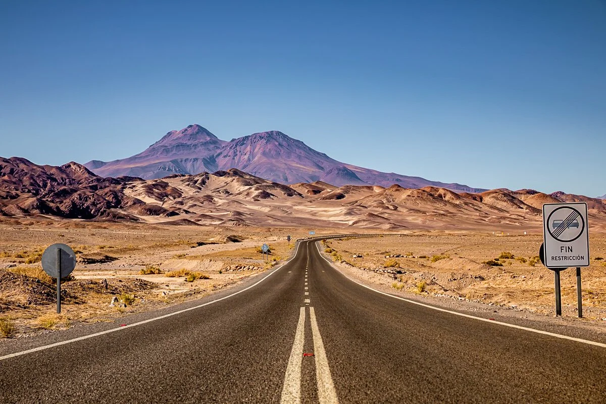A long, empty desert road stretching to distant mountains under a clear blue sky, with road signs at the roadside.