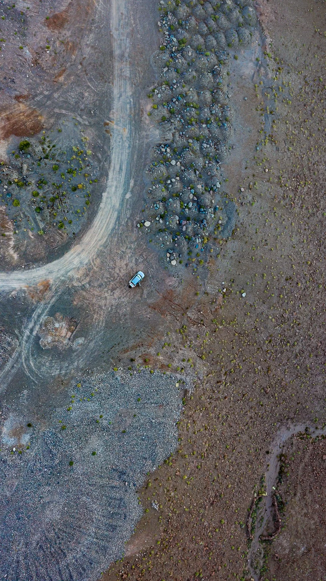 An aerial view of a dirt road winding through a rocky desert landscape with sparse green vegetation and a single white vehicle parked near the center.