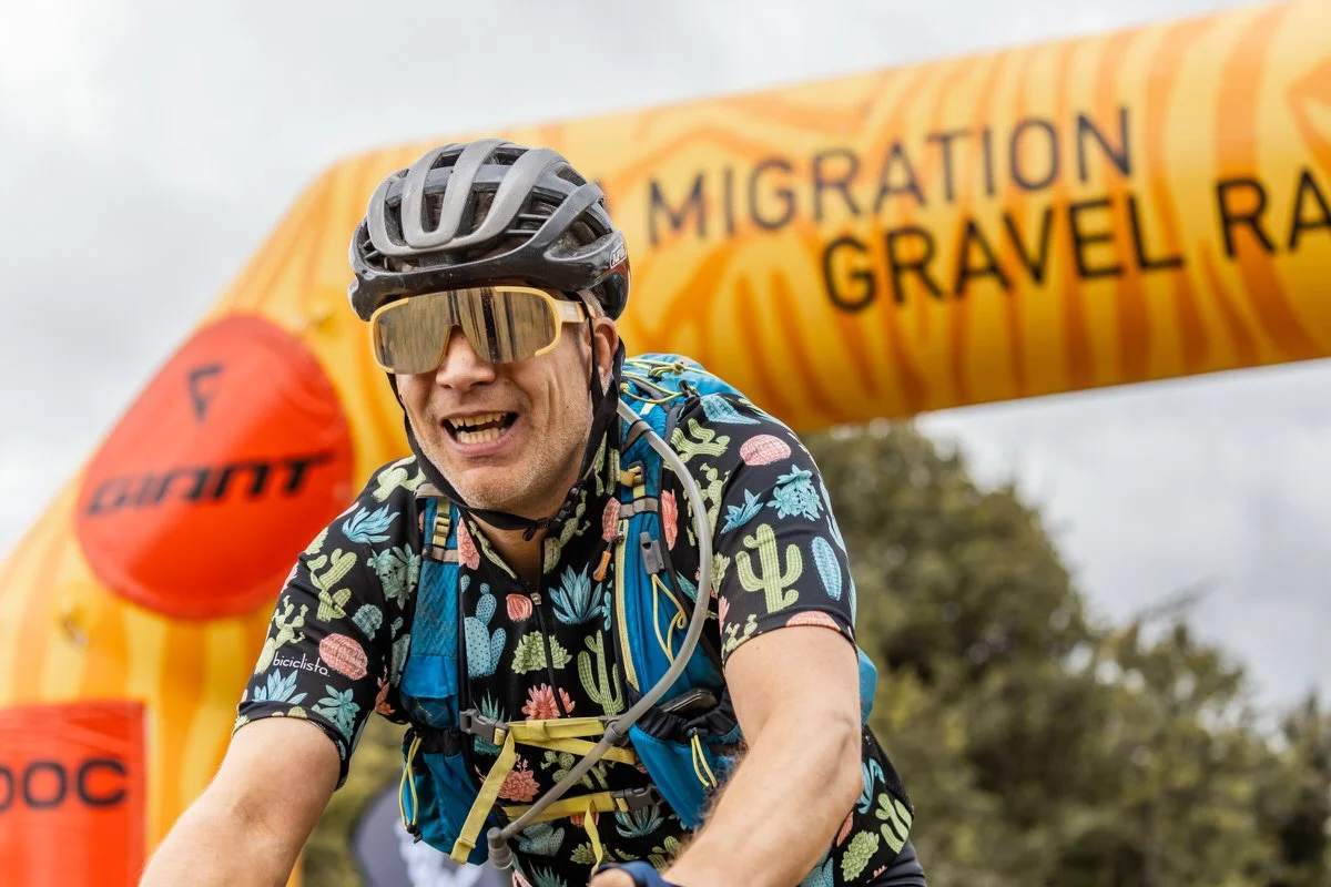 Male cyclist wearing a black helmet, reflective sunglasses, and a colorful floral shirt, riding during a race under a yellow inflatable arch with "Migration Gravel Rally" written on it.