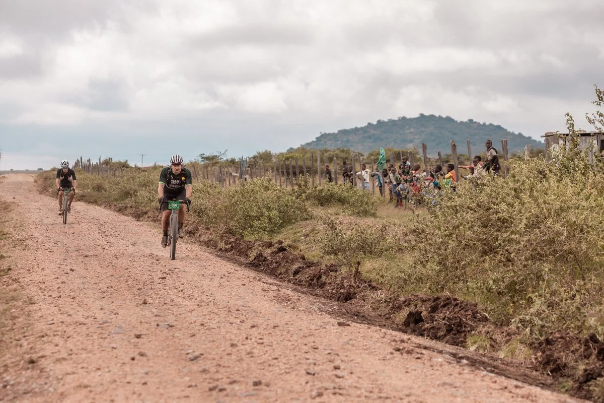 Two cyclists riding bikes on a dirt trail with a group of spectators behind a fence, mountain in the background, under cloudy sky.