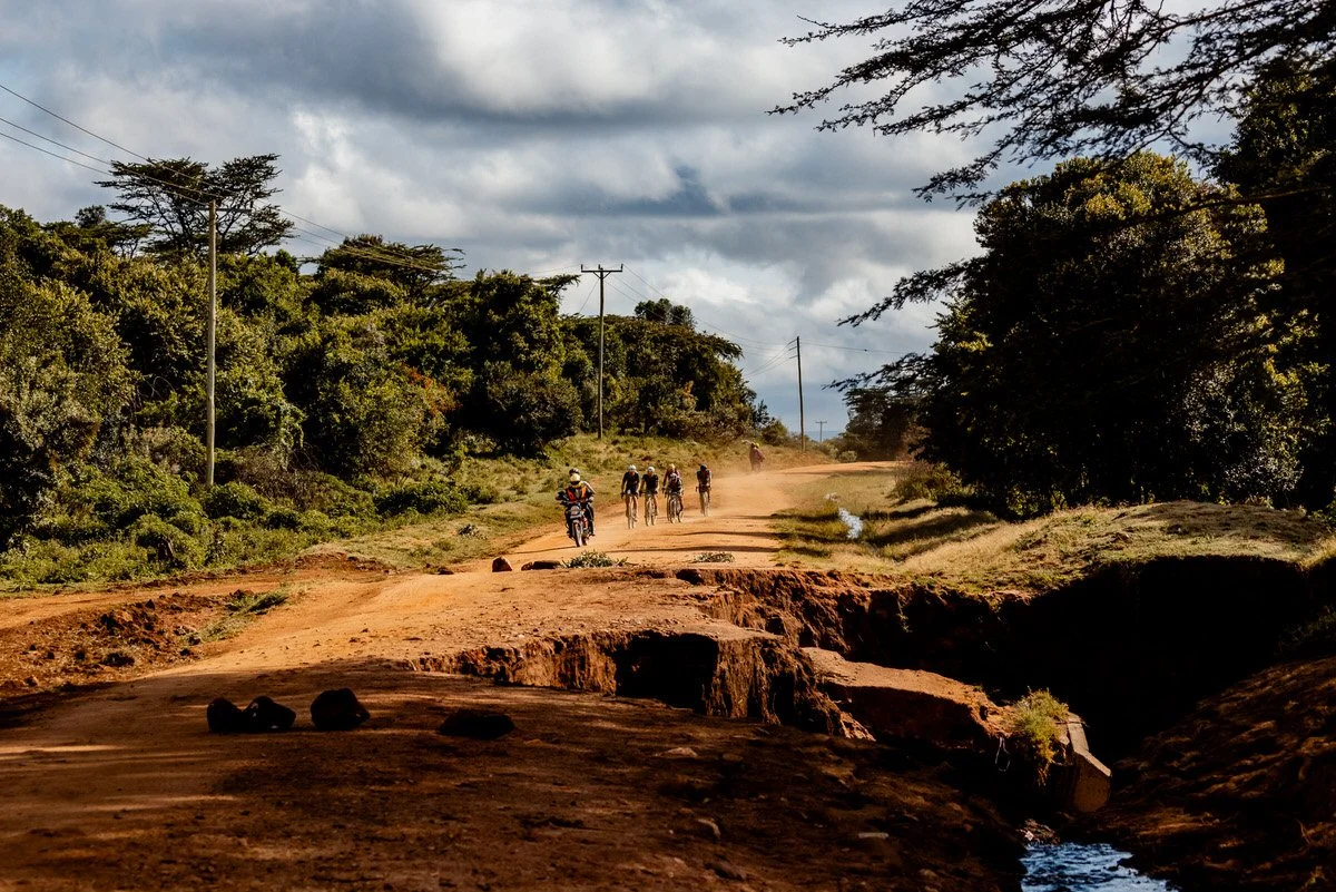 A dirt road with a group of cyclists and a motorbike riding along it, surrounded by trees and power lines under cloudy sky.