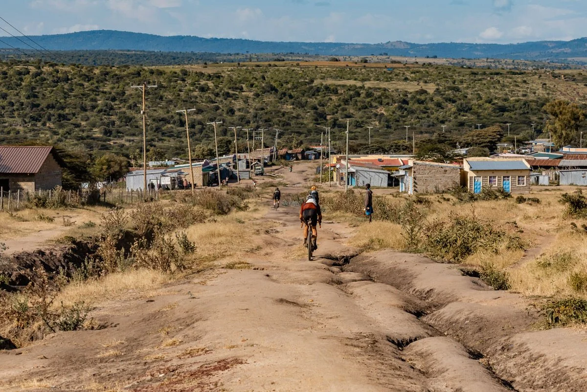 A dirt road leading to a small rural village with simple houses, a cyclist riding towards the village, and a few pedestrians walking along the road, with hills in the background under a partly cloudy sky.