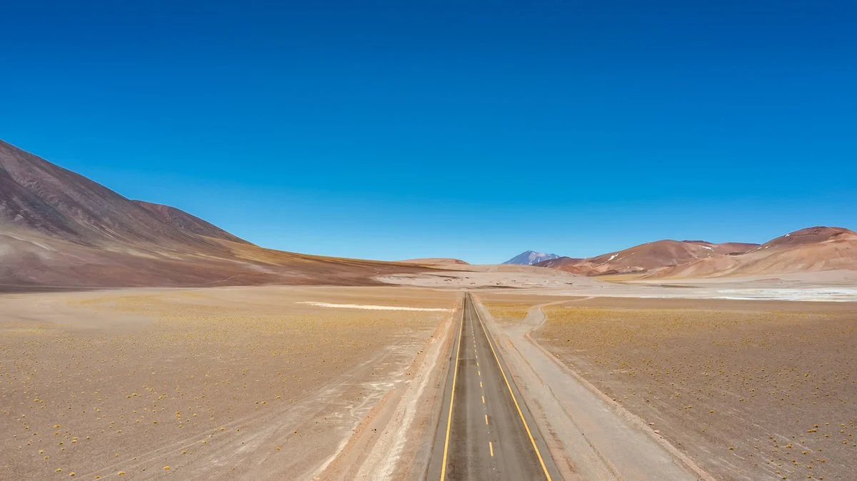 Long straight road through a barren desert landscape with mountains in the distance and a clear blue sky above.