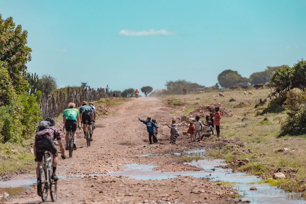 Group of children and adults standing and playing on a dirt road with puddles, while a few adults on bicycles ride away in the background in a rural area with green trees and grass.