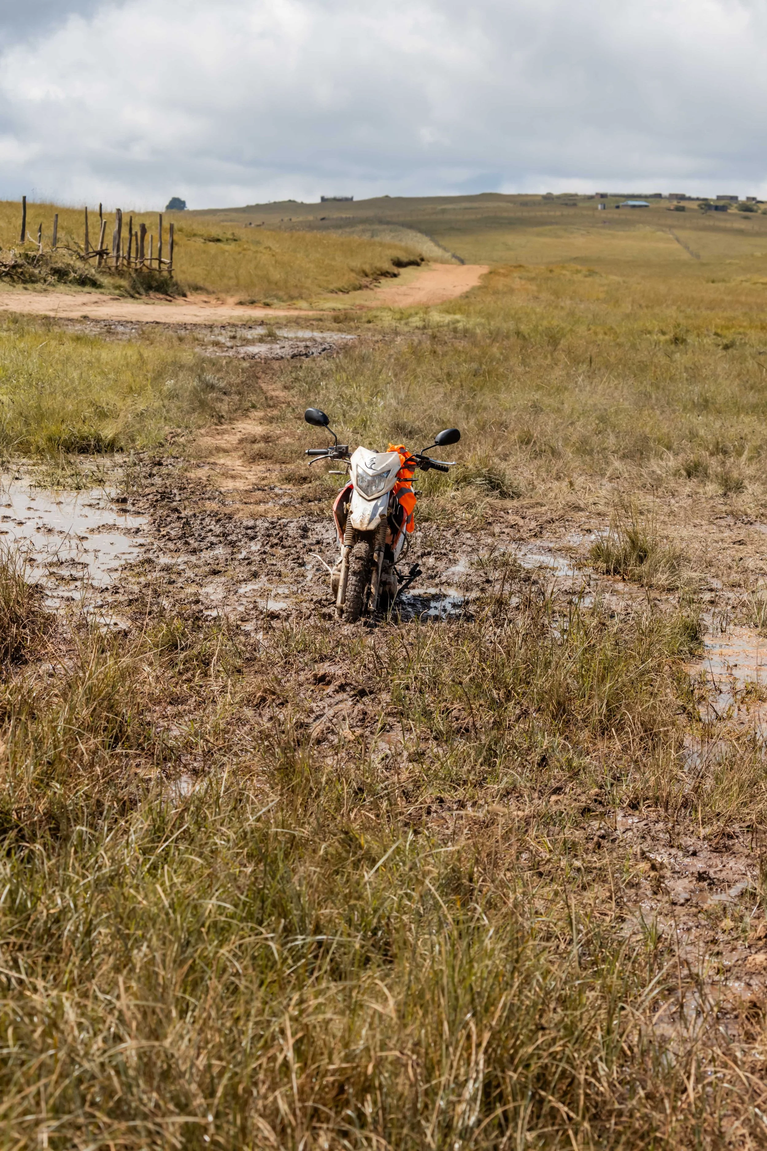 Motorcycle stuck in muddy field on a dirt road in a rural landscape with grassy hills and cloudy sky